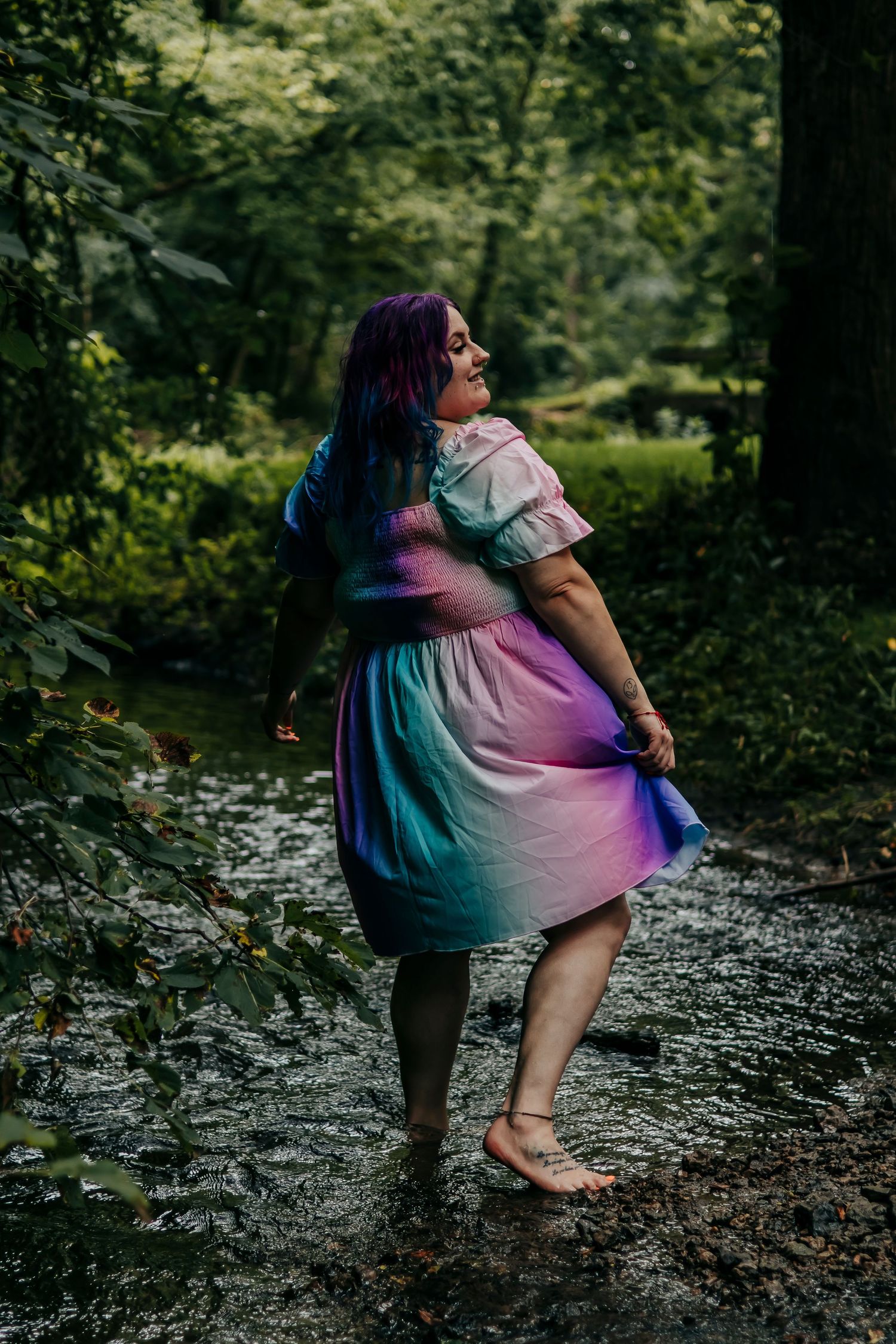 Person in colorful tie-dye dress twirls playfully in forest creek setting.
