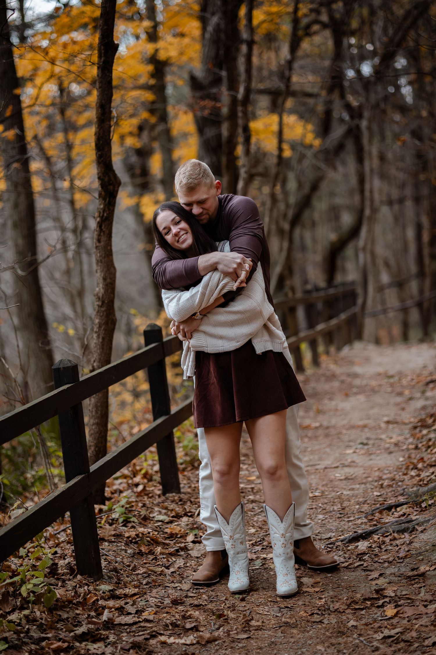 A couple embraces on a wooded trail surrounded by autumn trees with golden leaves and a wooden railing.