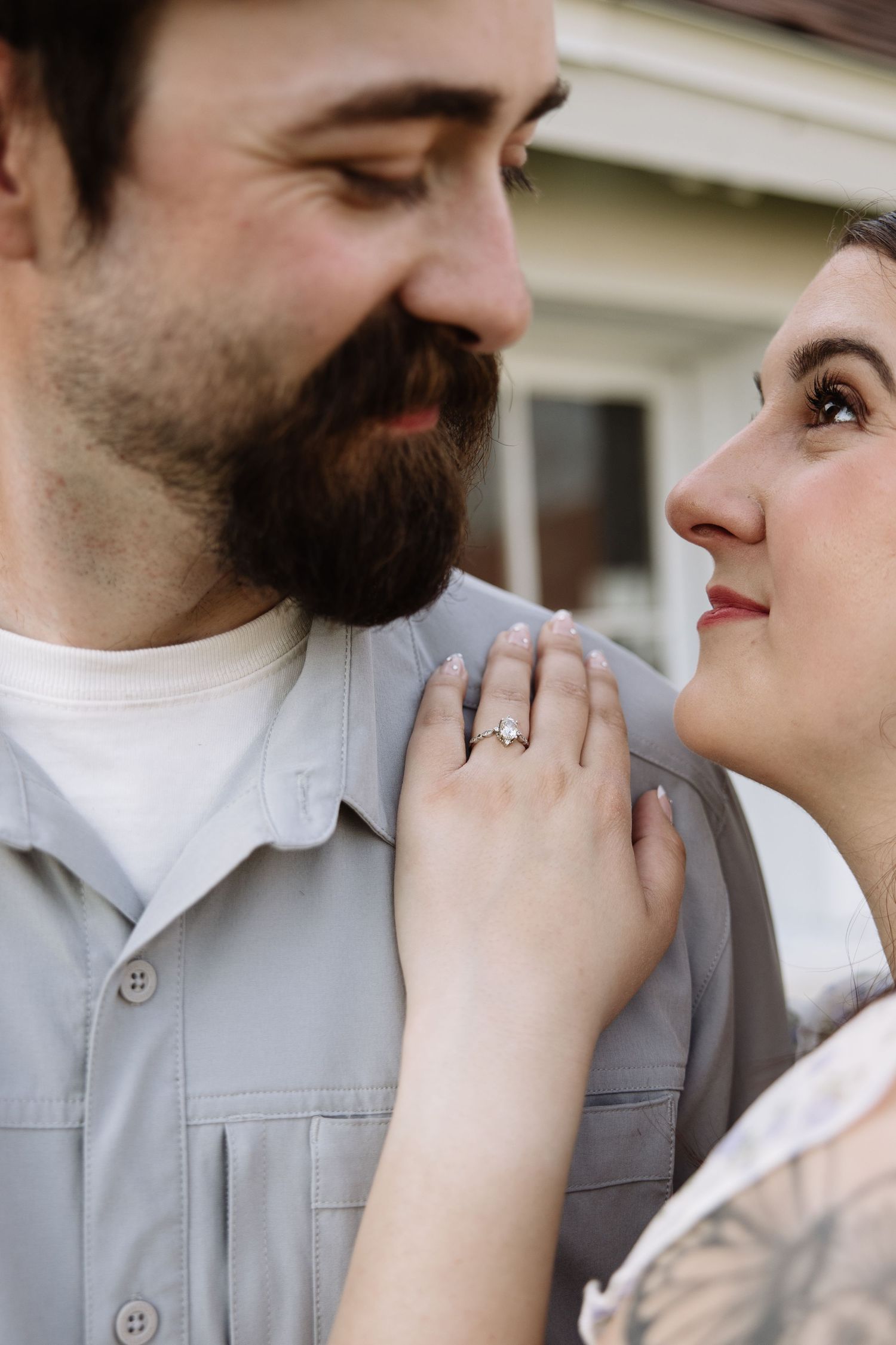 A bearded man and woman share a tender moment, her tattooed hand displaying a diamond engagement ring on his shoulder.