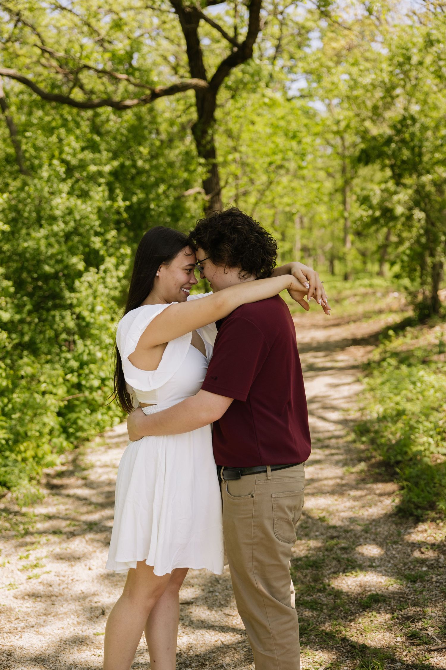 Happy couple hugging and touching foreheads on a sunlit forest path, woman in white dress and man in red shirt.