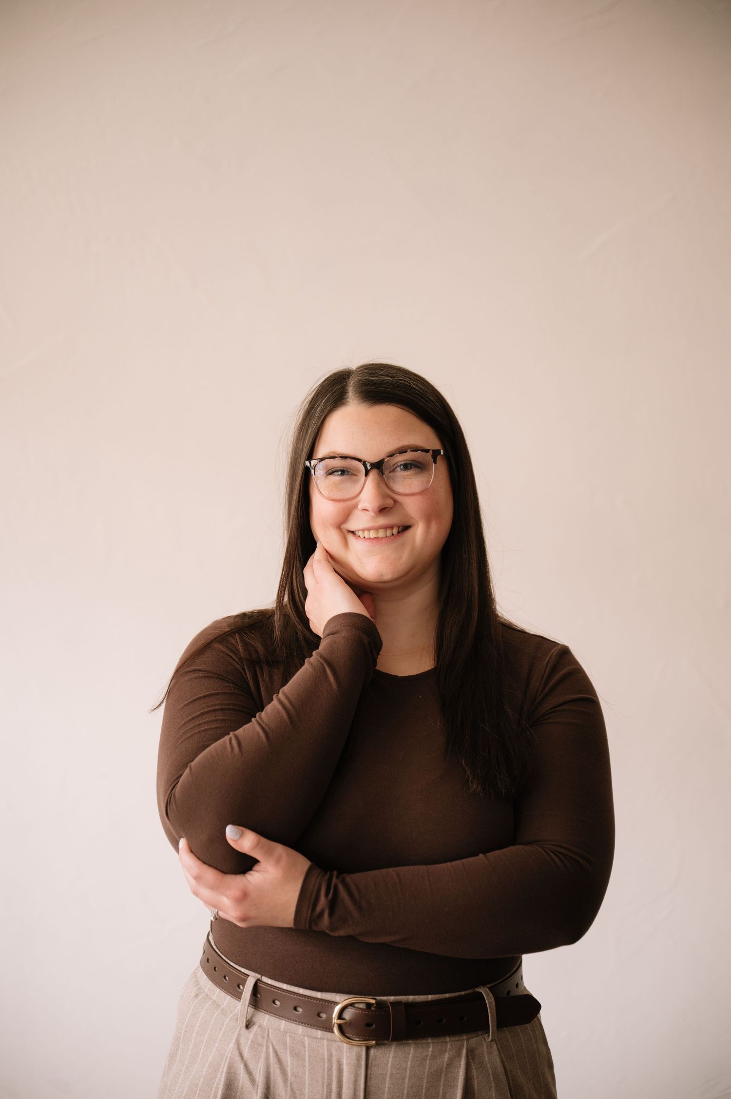 A person wearing glasses and a brown sweater poses with hand on chin against a light colored wall.