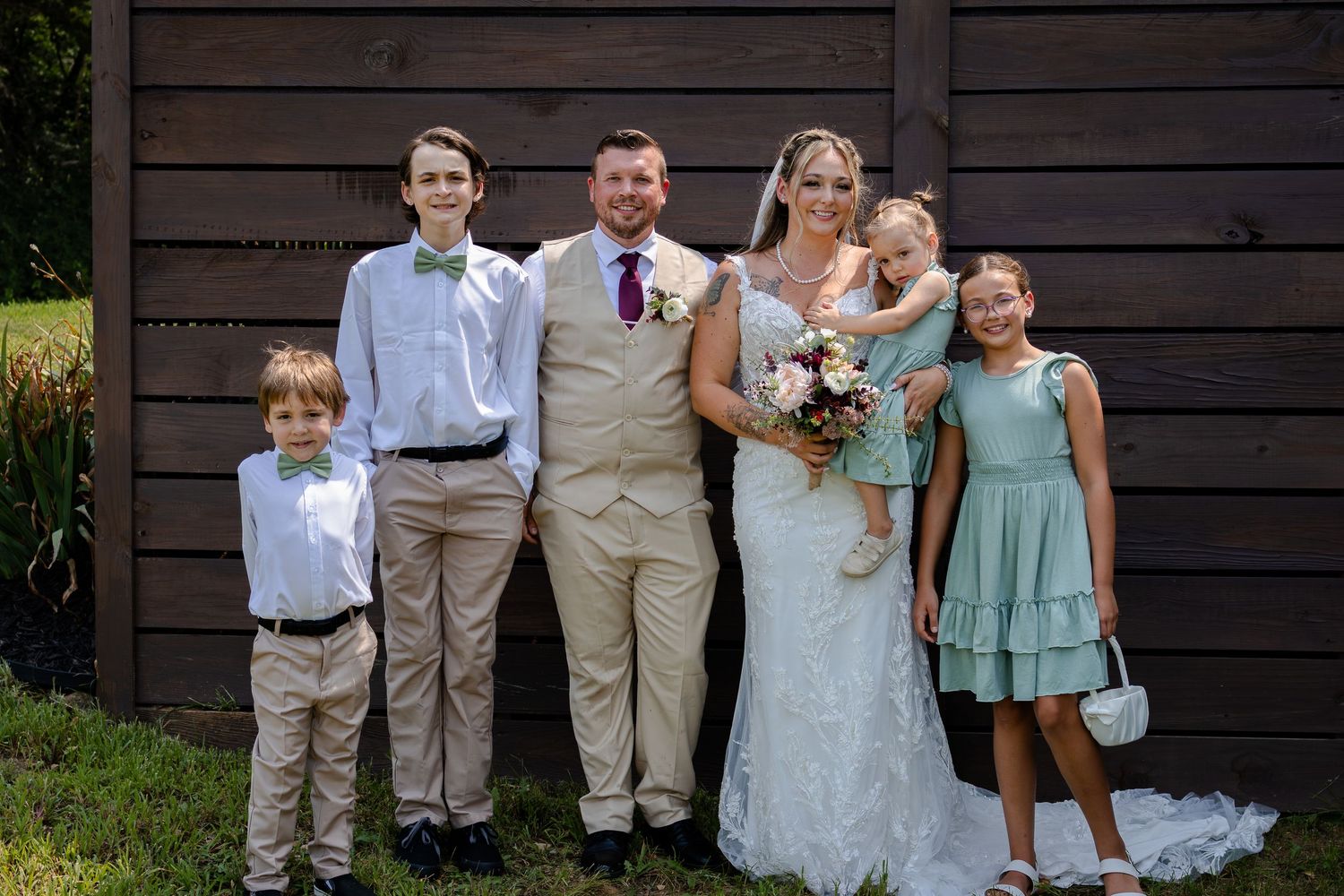 Wedding party portrait of bride and groom with four children in formal attire posing outdoors near a wooden barn.