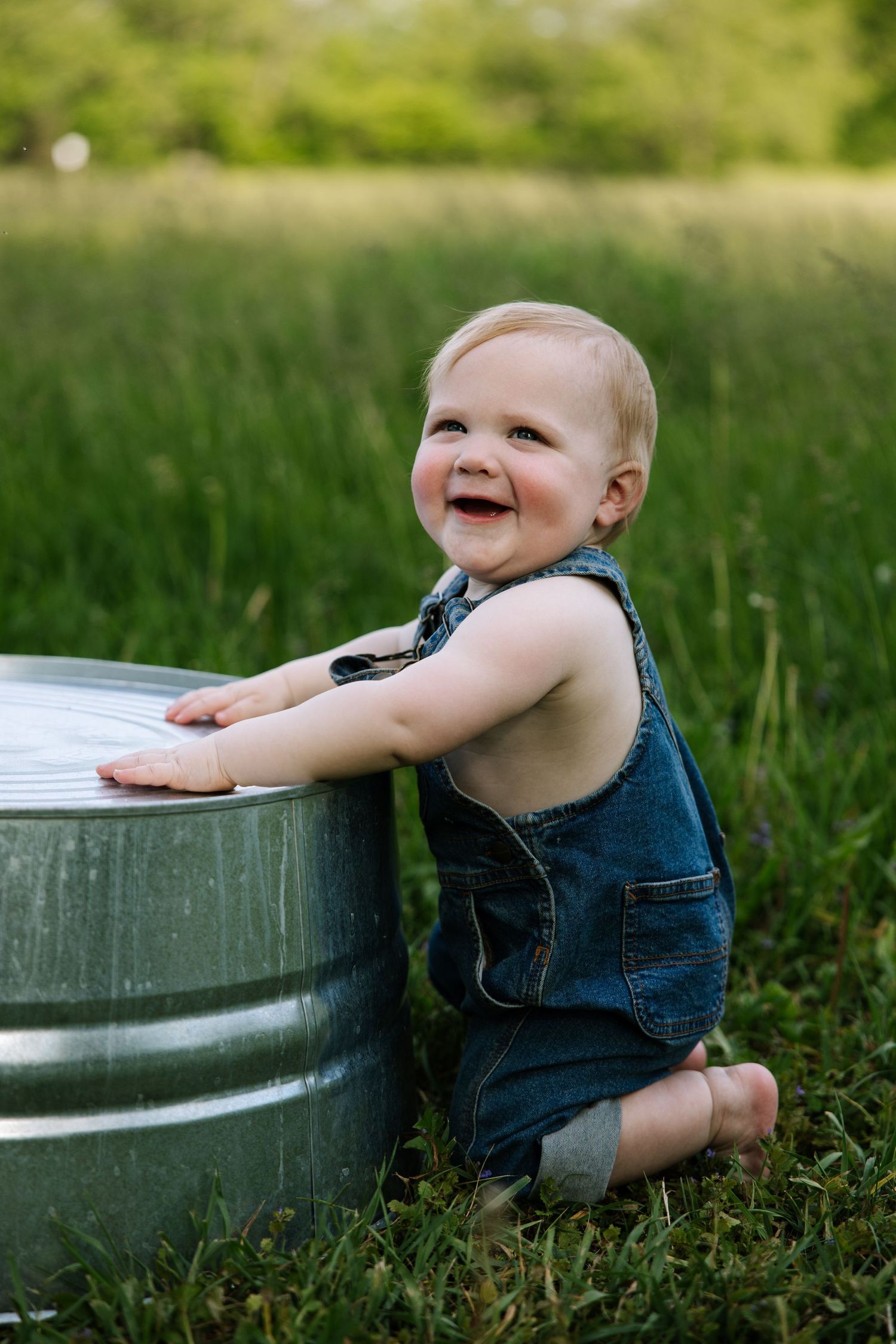 Smiling baby in denim overalls kneeling on grass beside a galvanized metal tub outdoors.