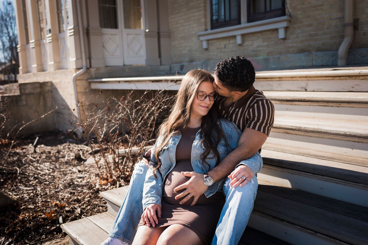 Couple sitting on steps outdoors, man embracing pregnant woman from behind, both looking at her baby bump.