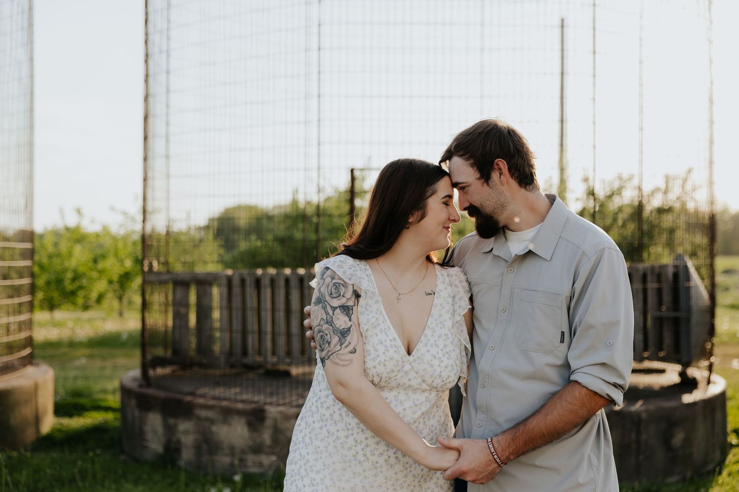 A couple shares a tender forehead-to-forehead moment in a sunny farm setting, dressed in casual elegant attire.