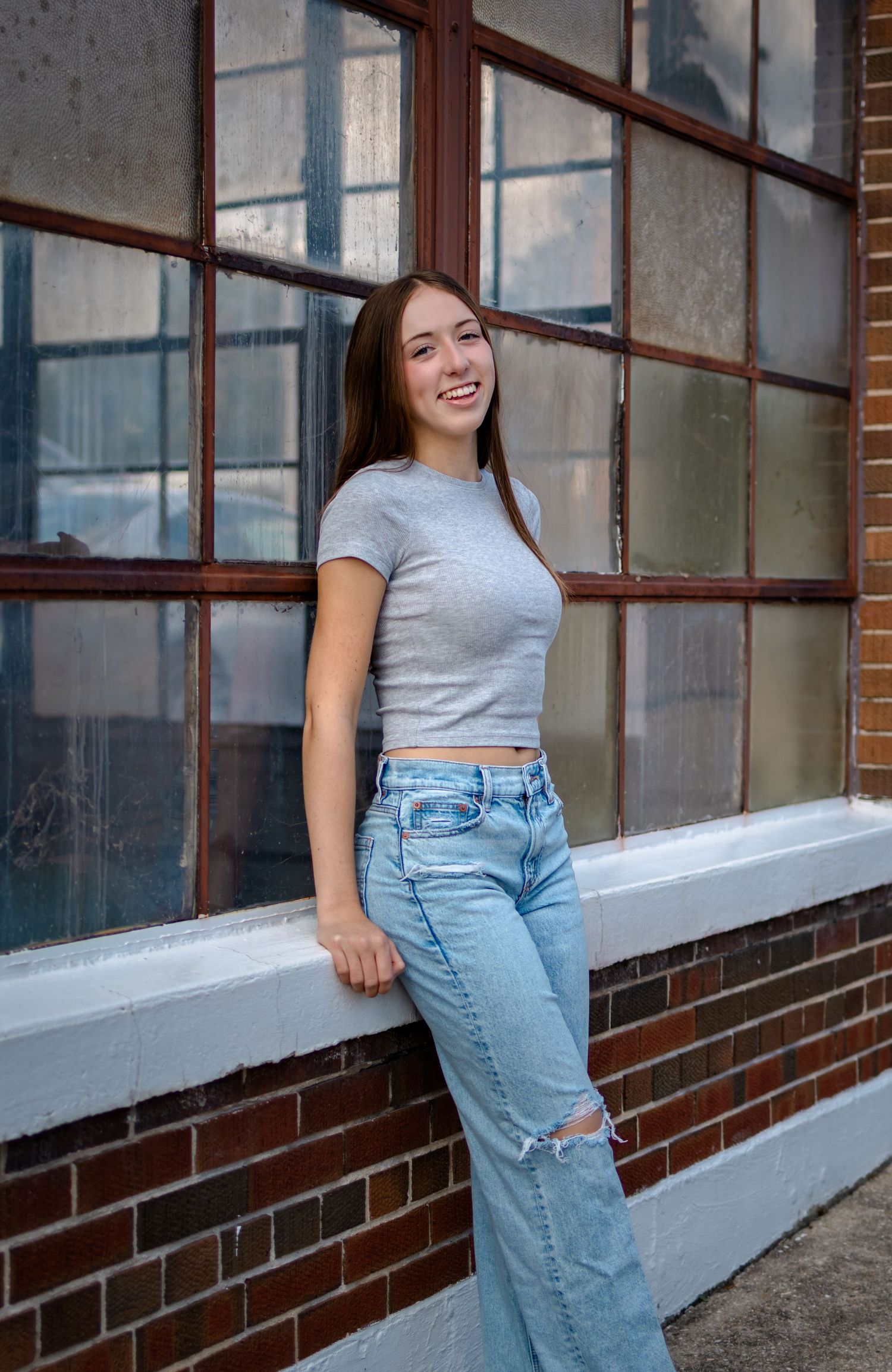 A person wearing a gray t-shirt and light blue ripped jeans poses casually against a brick building exterior.