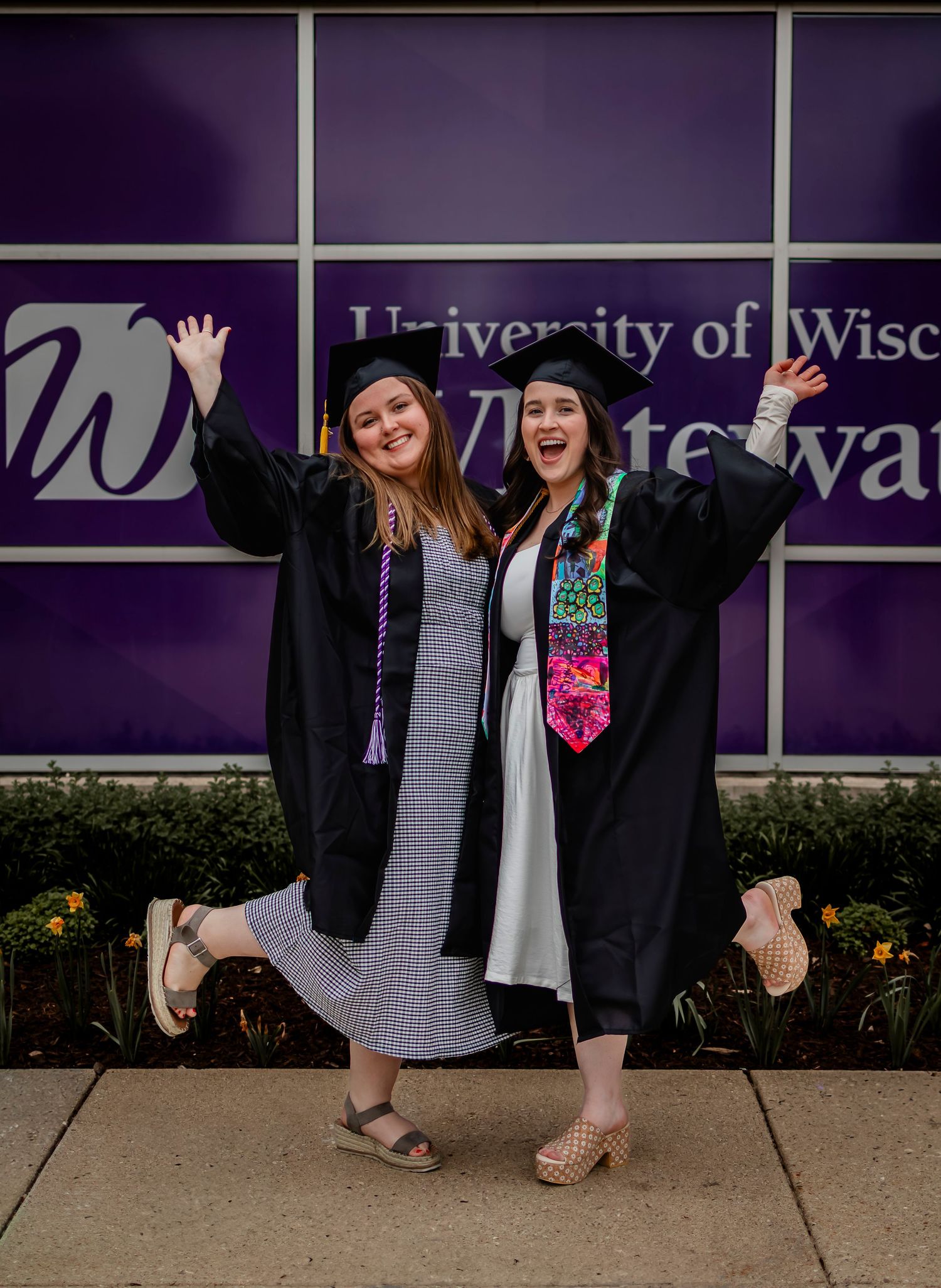 Two graduates in black caps and gowns celebrate joyfully outside University of Wisconsin-Whitewater.
