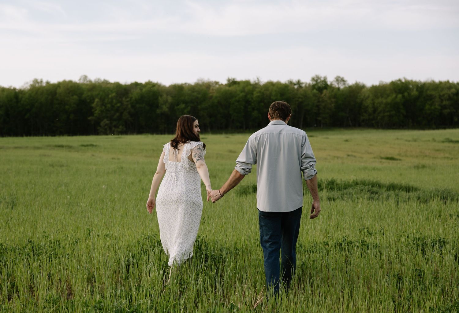 Couple holding hands walking through lush green field, woman in white dress, man in blue shirt, trees in background.