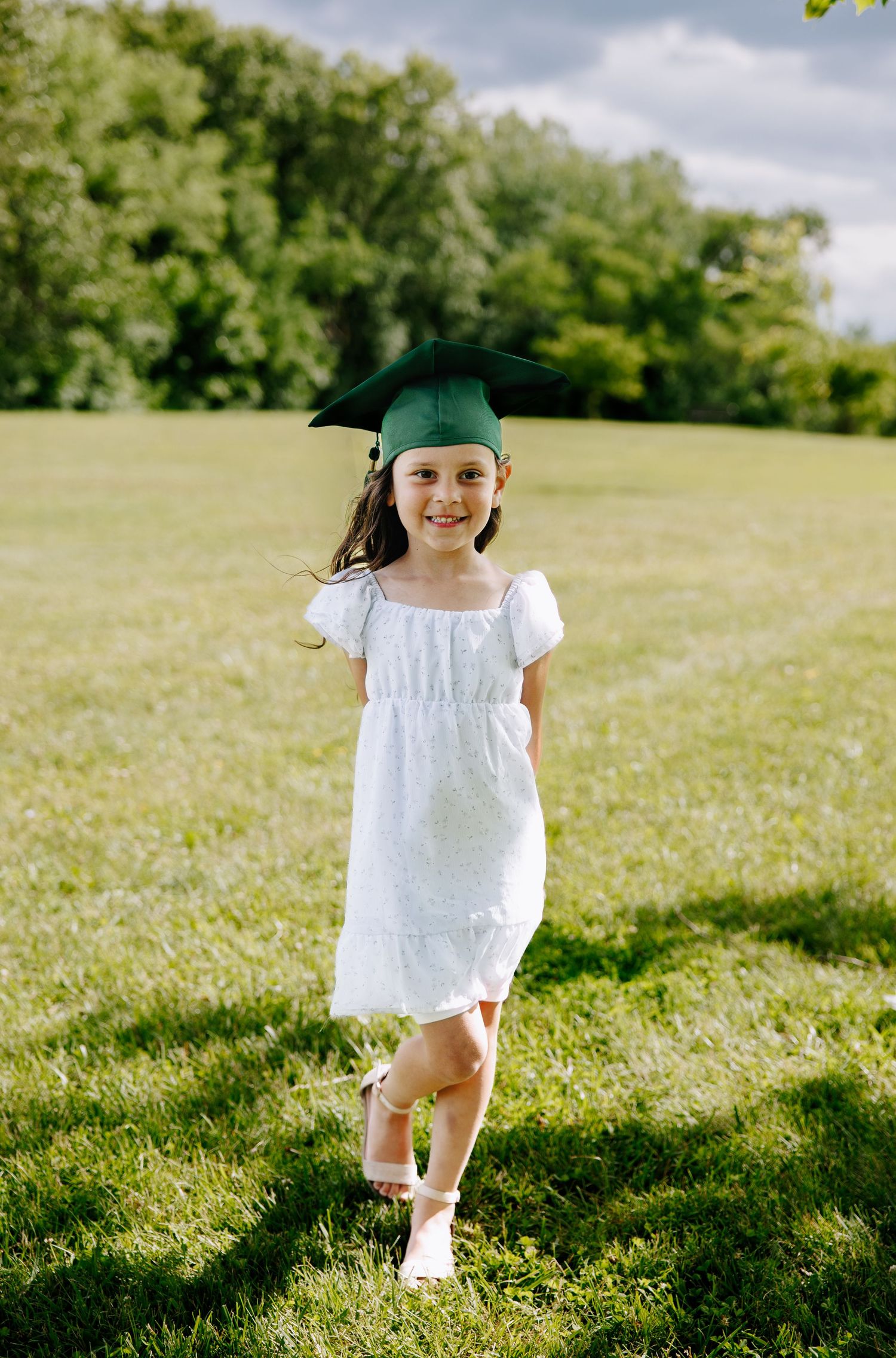 Young girl in white dress and green graduation cap smiling outdoors on green grass field.