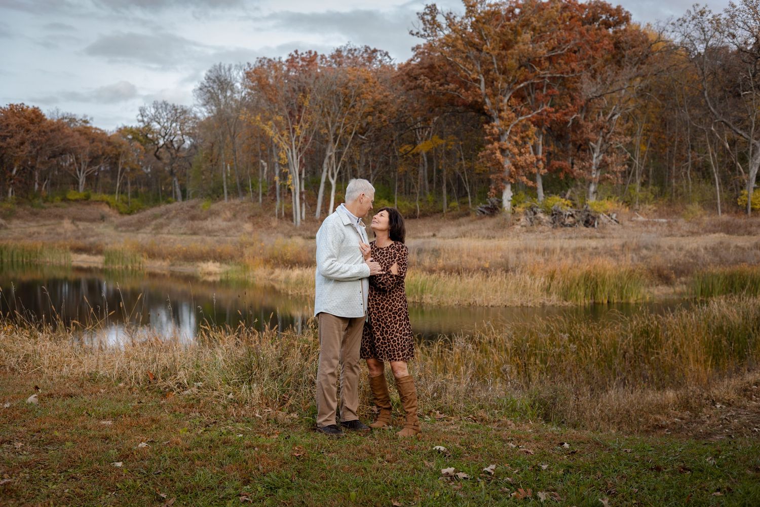A couple strolls through an autumn landscape with tall trees and golden grass near a tranquil pond.