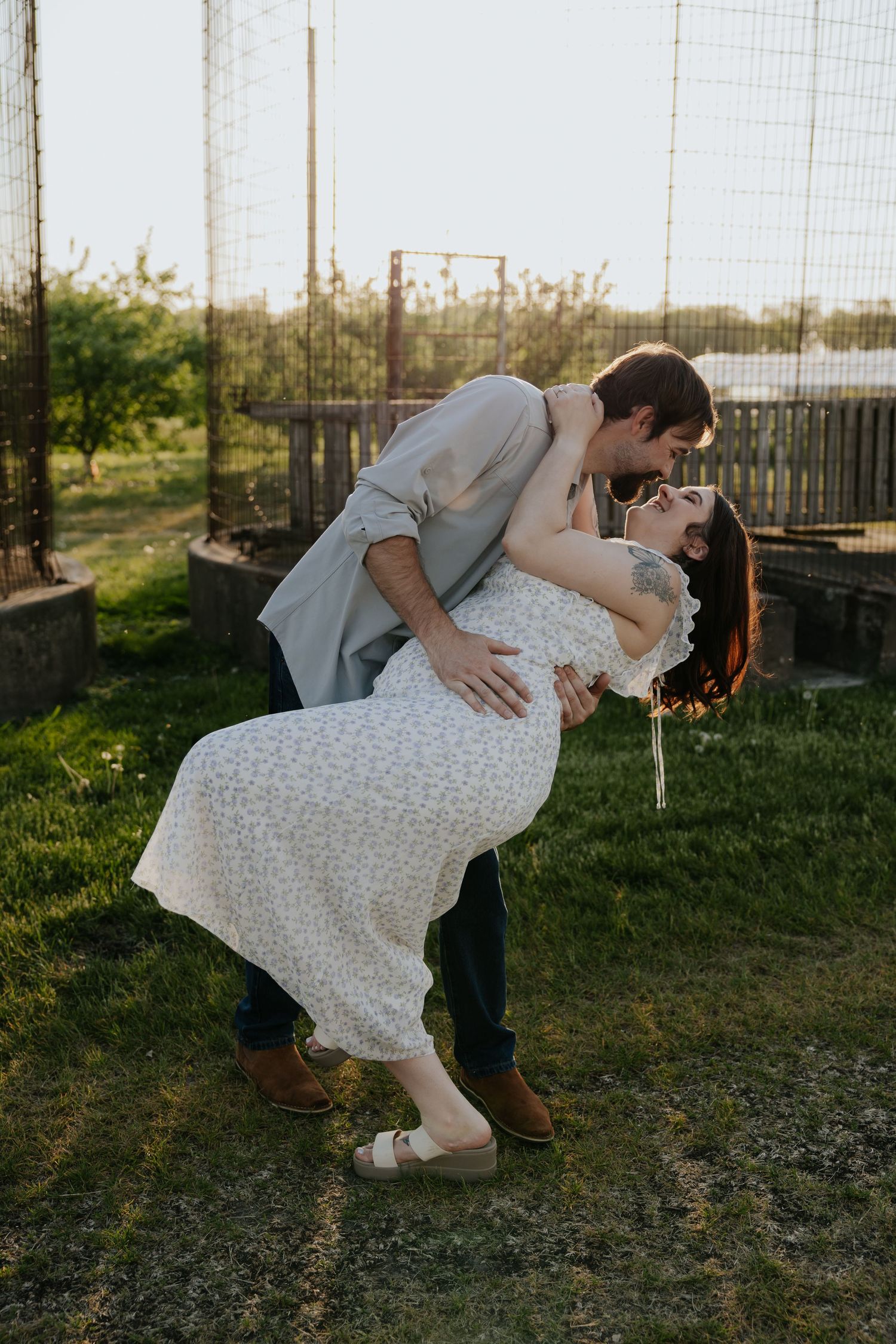 A couple shares a romantic dip kiss at golden hour in a lush garden setting, the woman in a floral dress.