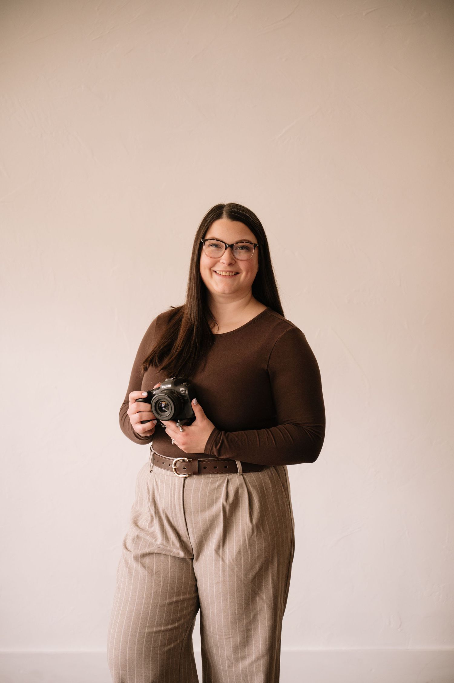 Professional photographer wearing brown sweater and striped pants holds camera in studio setting.