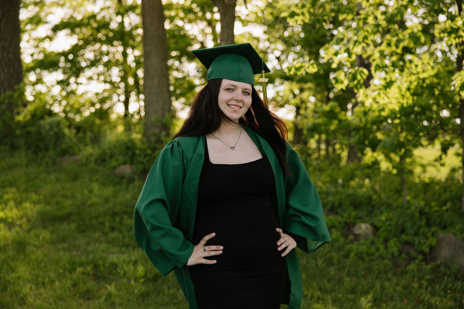 Young woman in green graduation cap and gown smiling outdoors in lush green park setting.
