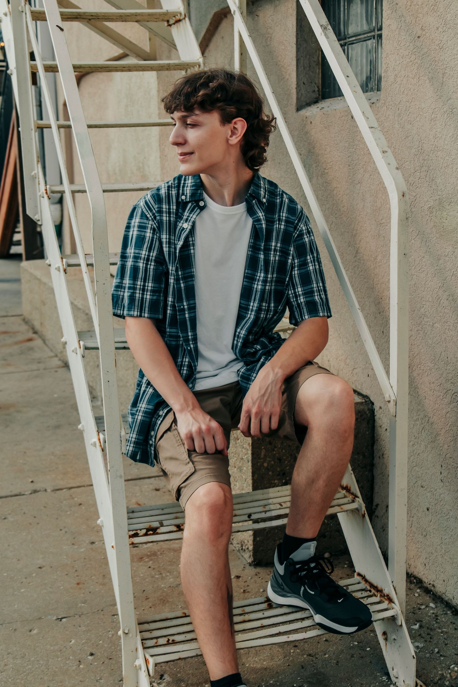 Young person in plaid shirt and shorts sits casually on outdoor metal stairs.