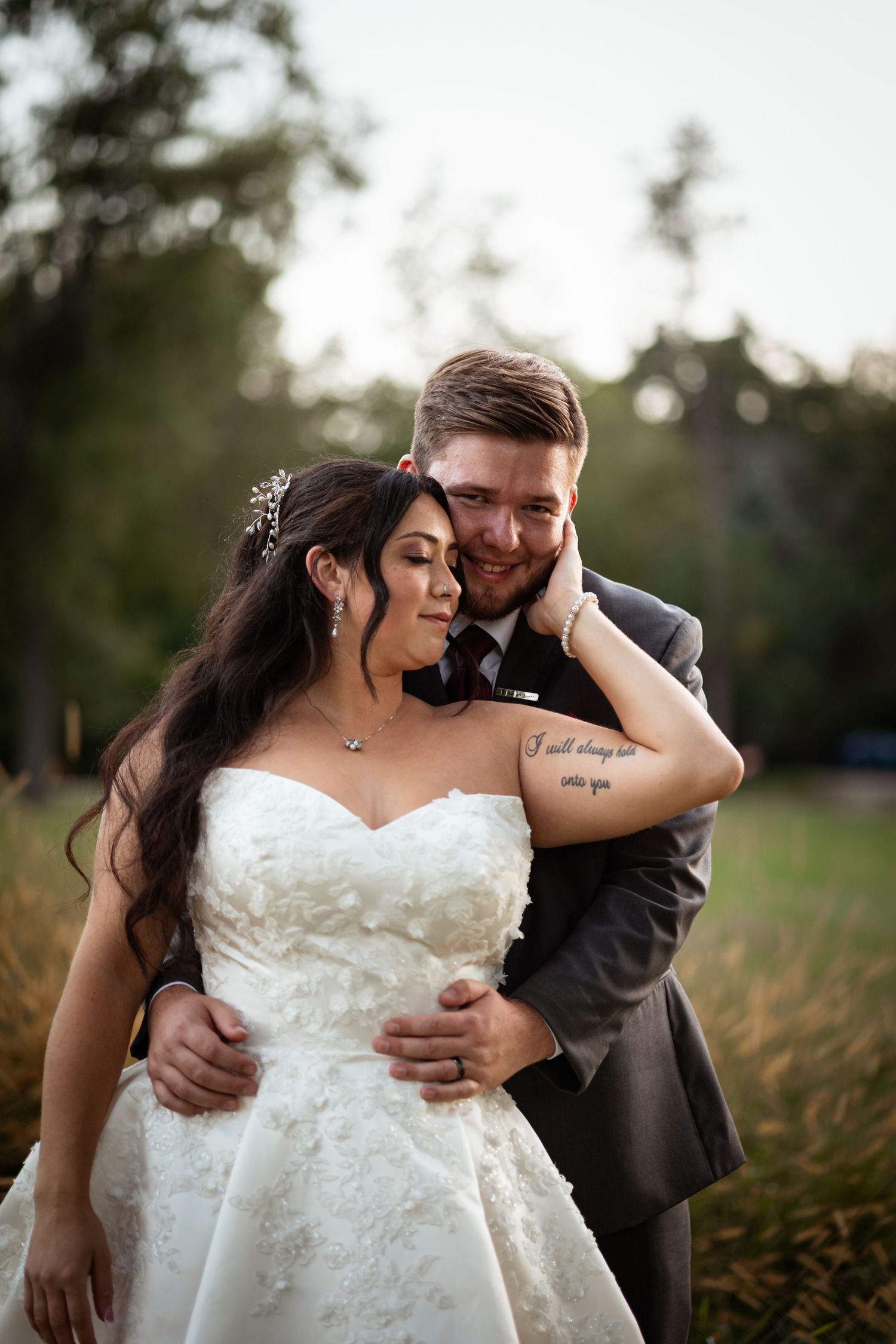A romantic wedding couple embraces in an outdoor setting with natural lighting and tall grass.