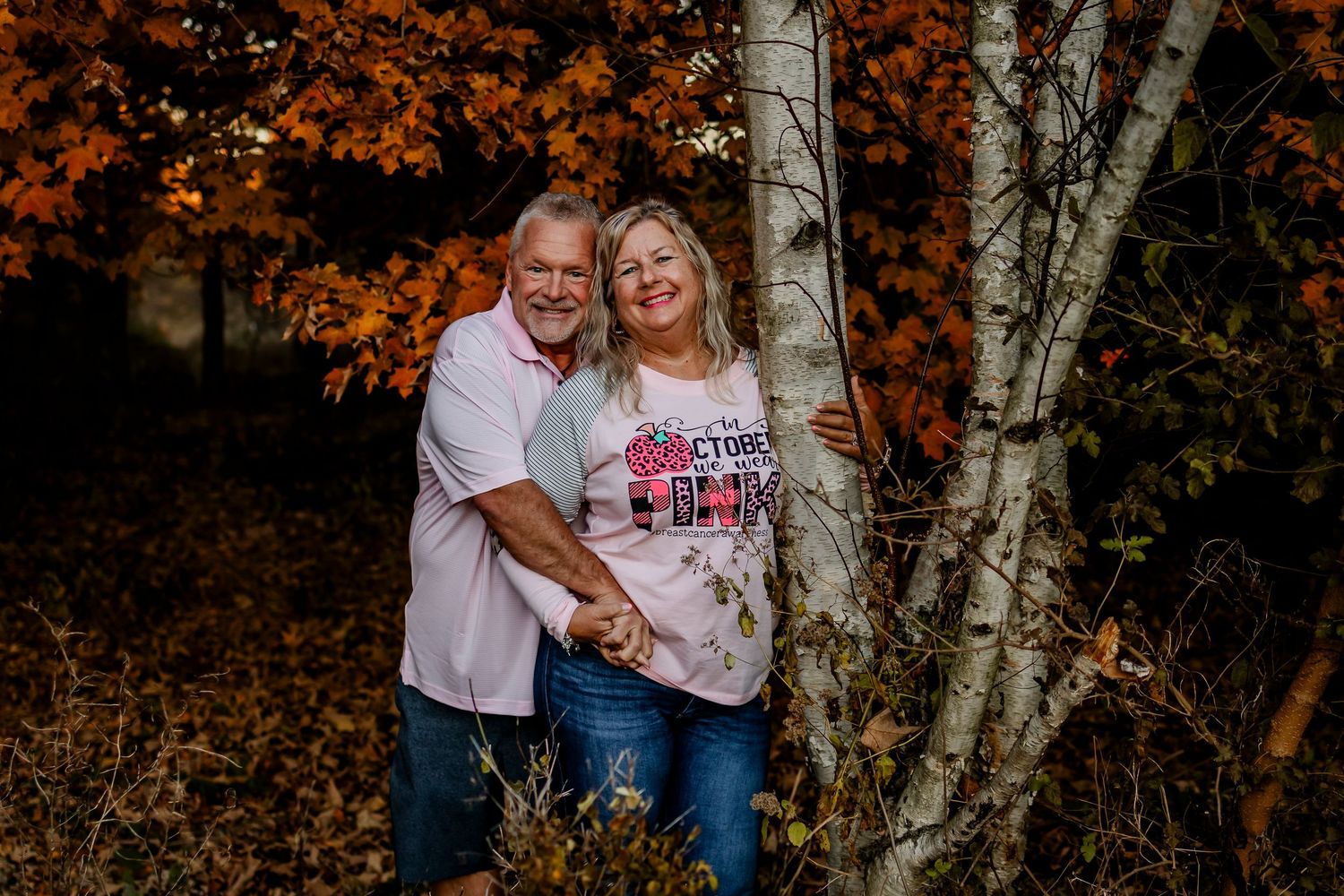 Couple embracing by white birch tree surrounded by vibrant orange autumn foliage in a fall outdoor portrait.