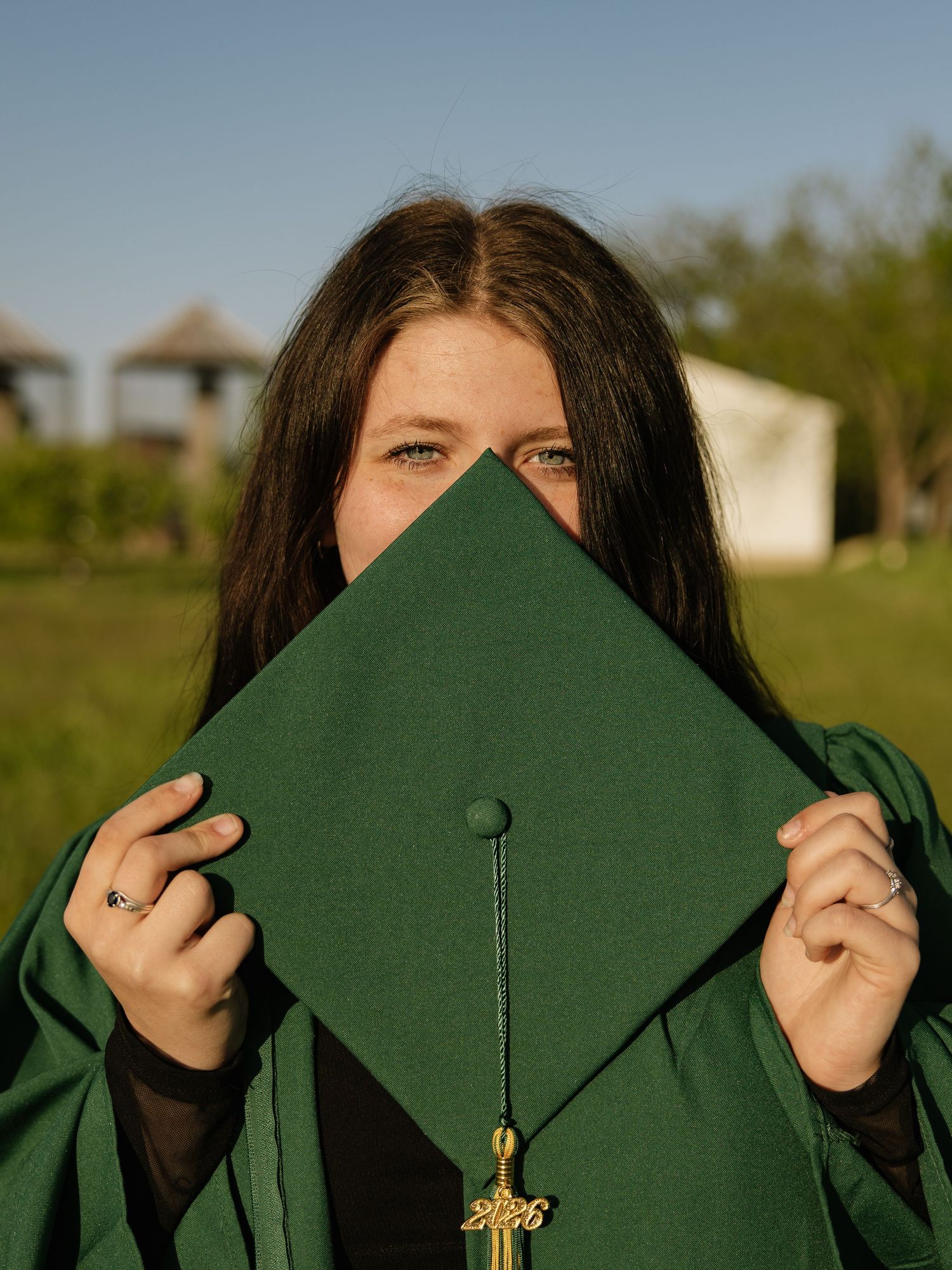 Graduate hiding face behind green mortarboard cap with gold tassel outdoors.