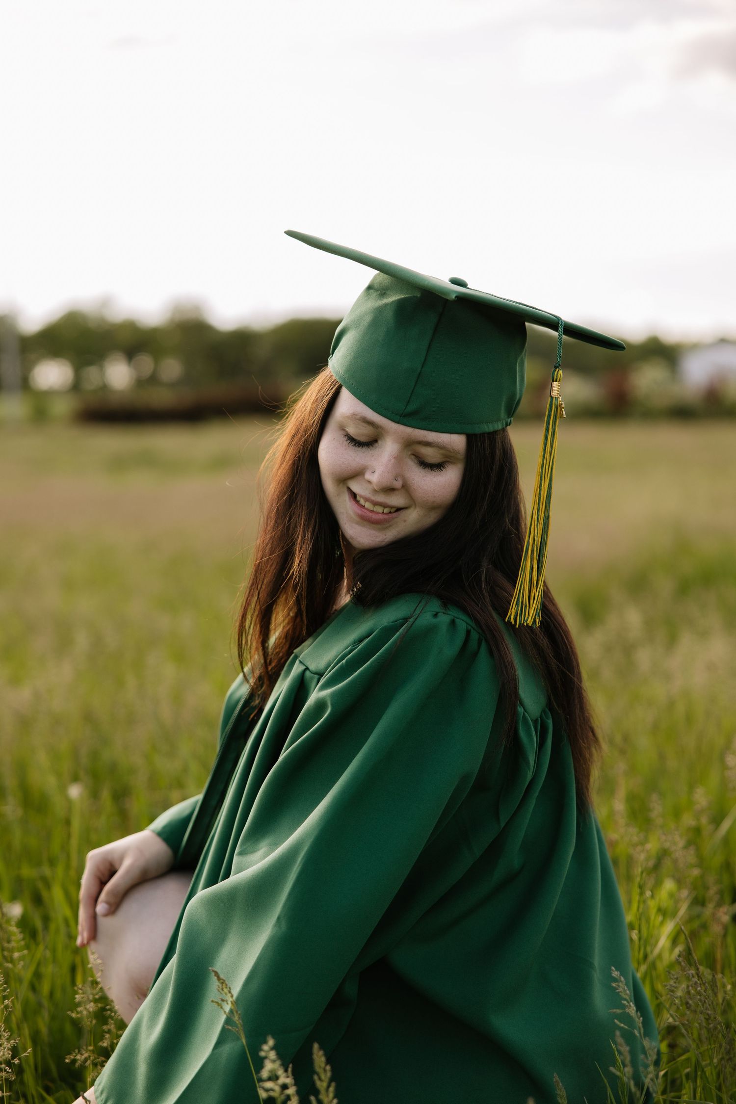 Young woman smiling in green graduation cap and gown, posing outdoors in a lush green field.