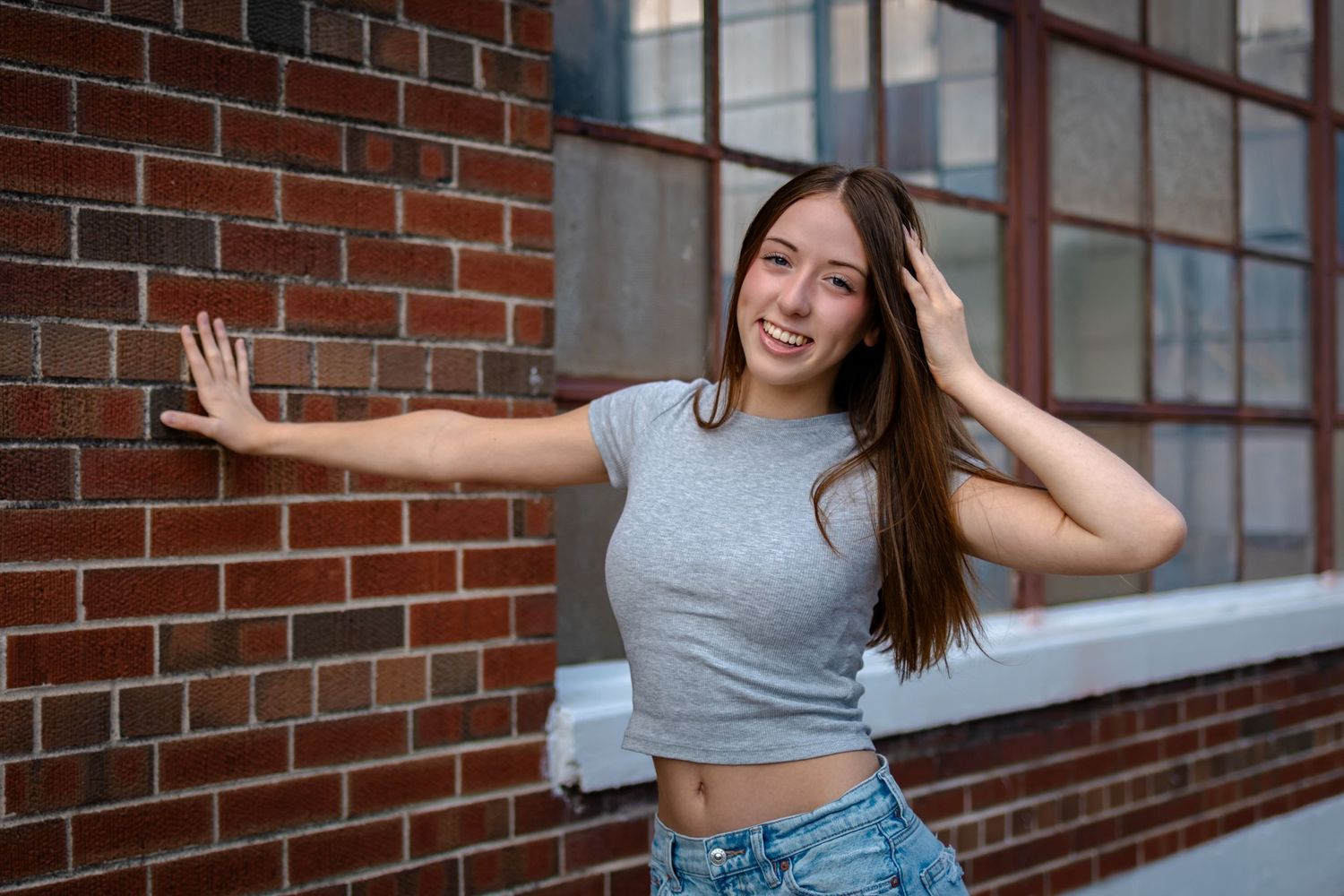 Woman in crop top and jeans poses playfully against brick wall background.