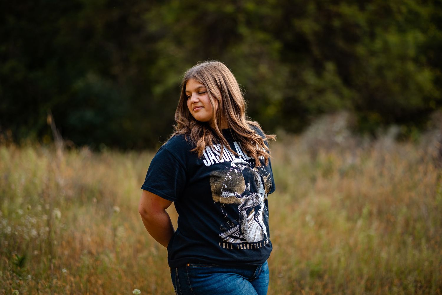 Person in a navy blue t-shirt walking through a sunlit meadow with tall grass and trees in the background.