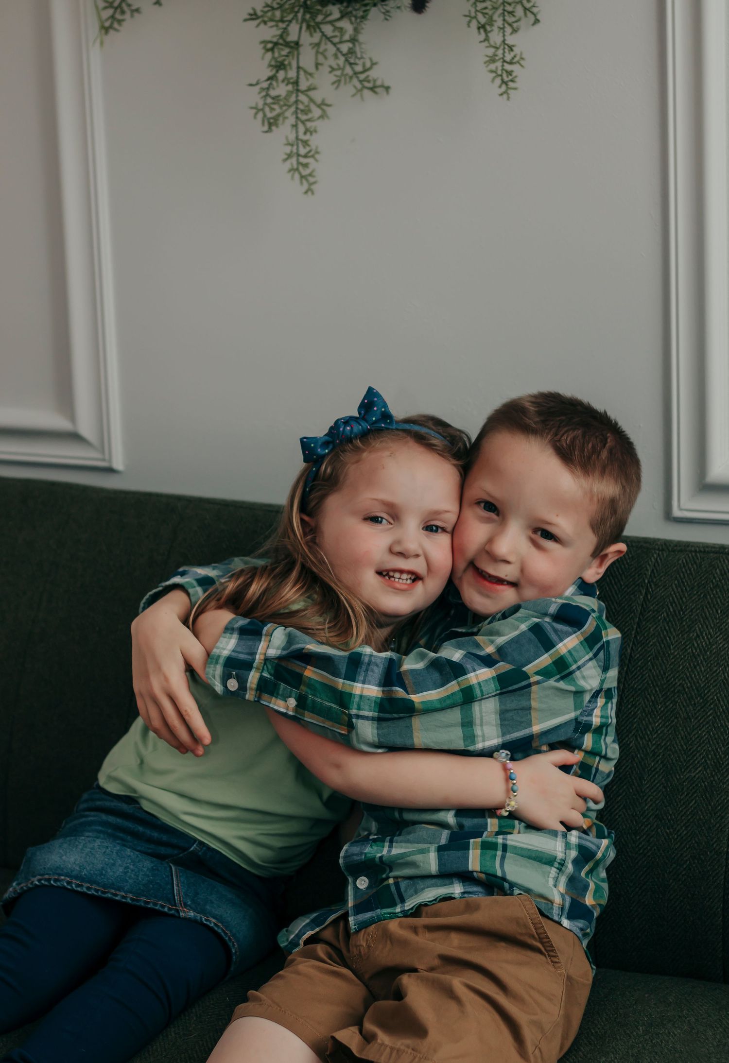 Two children sharing an affectionate hug while sitting on a dark green couch under decorative greenery.