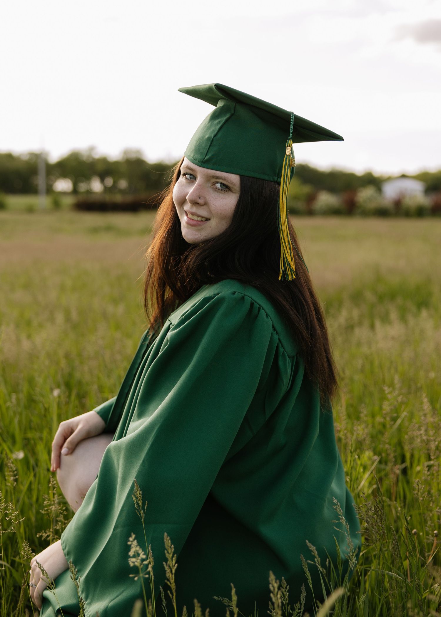 Young woman in green graduation cap and gown smiling in a field.