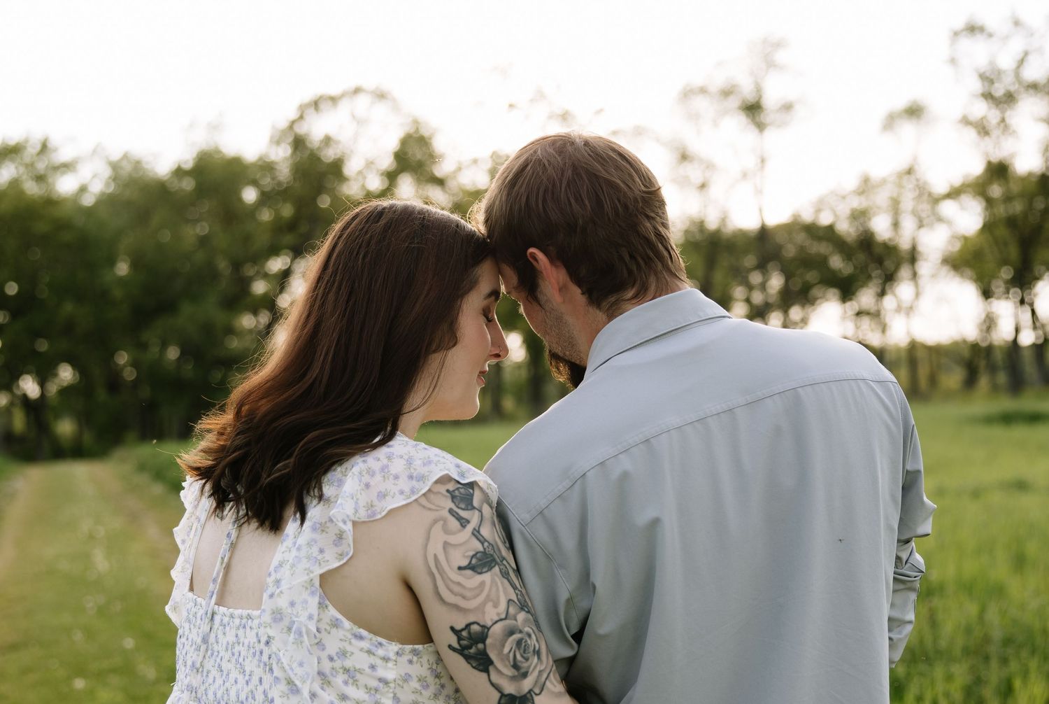 Couple sharing an intimate moment outdoors, woman with floral arm tattoo in lace top, man in light blue shirt, green field background.