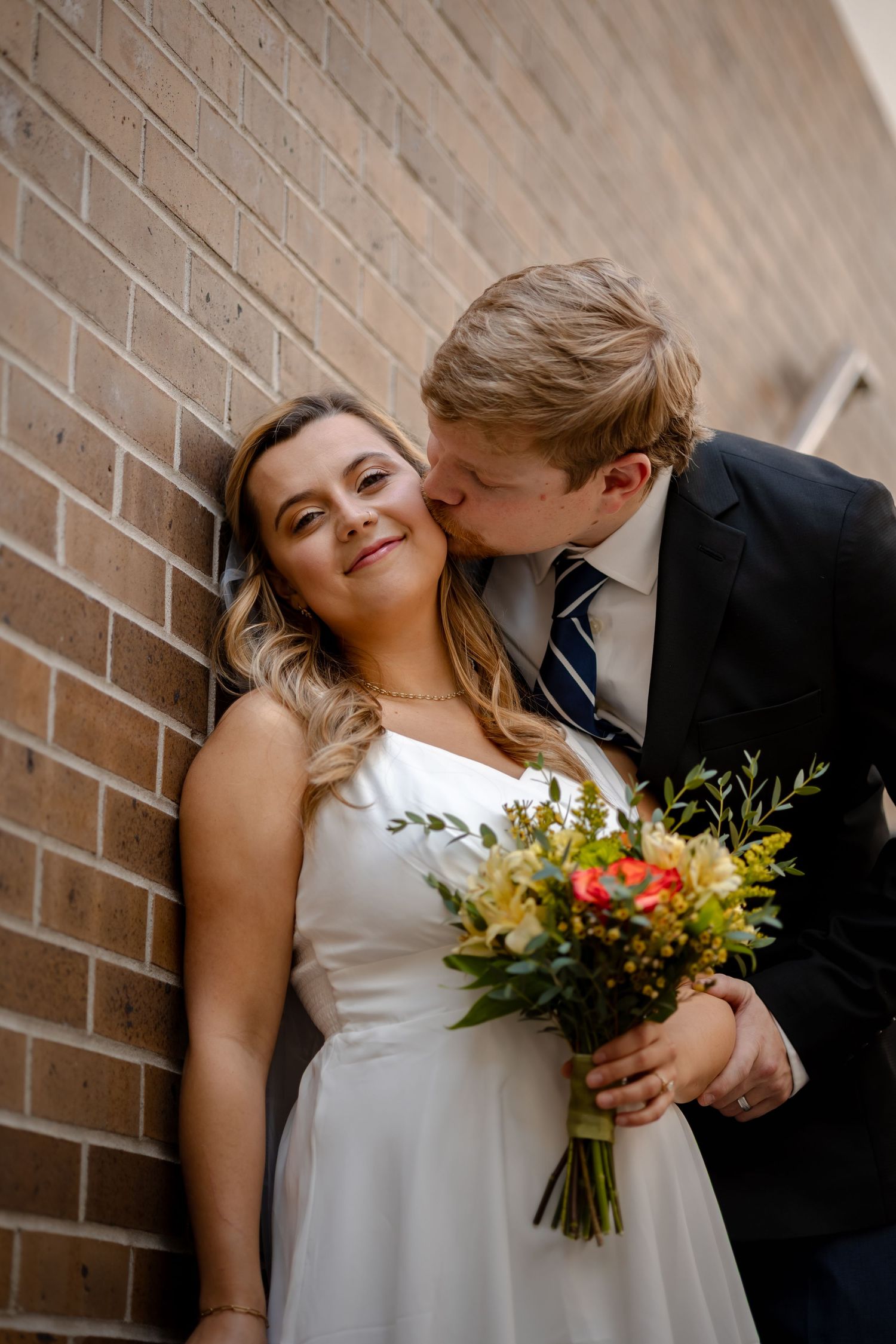 A groom in a navy suit kisses his bride on the cheek as she smiles holding a colorful bouquet against a brick wall.