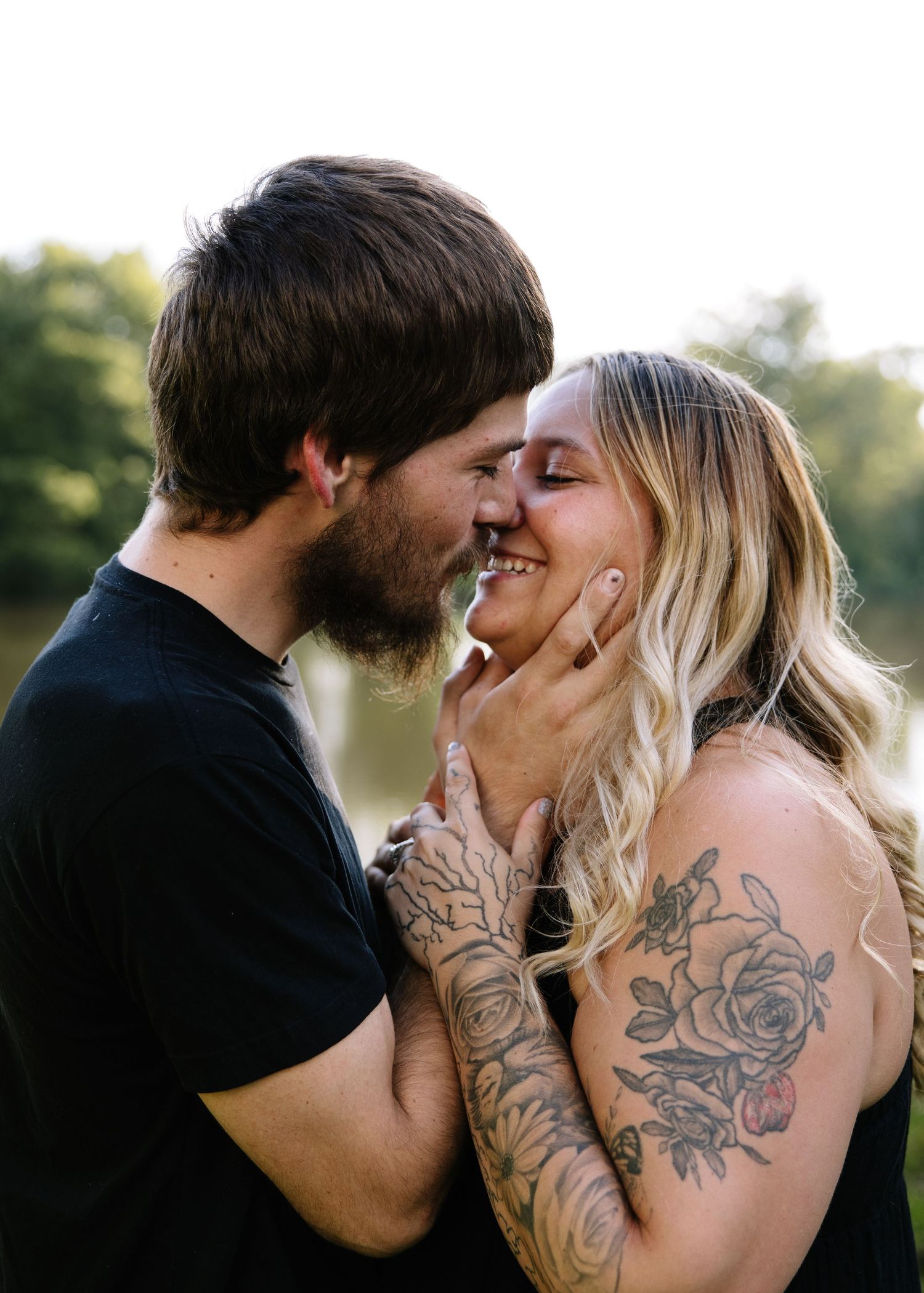 A tattooed couple shares a romantic almost-kiss outdoors near a lake, the man gently holding the smiling woman's face.