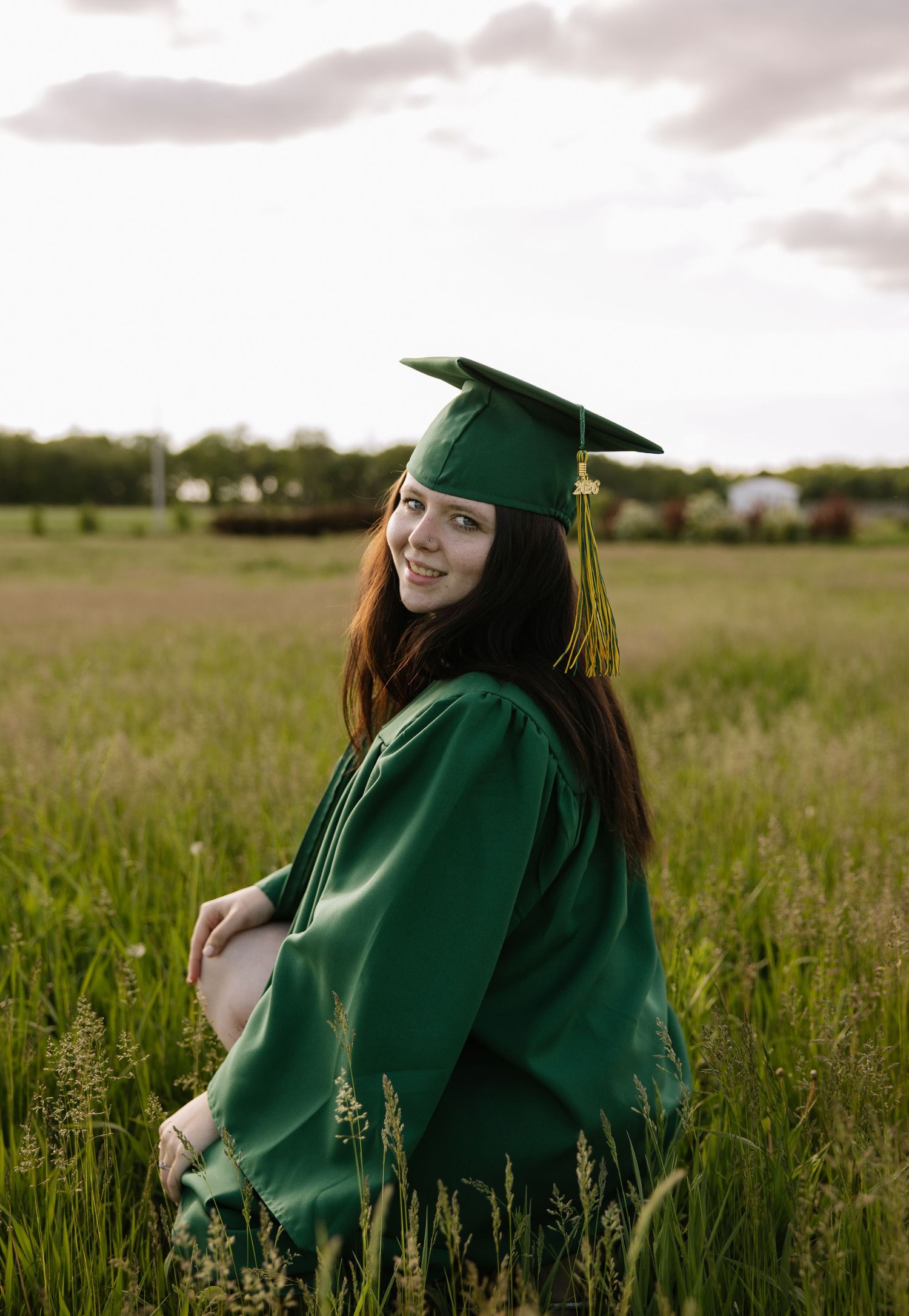 Young woman smiling in green graduation cap and gown standing in a lush green field at sunset.
