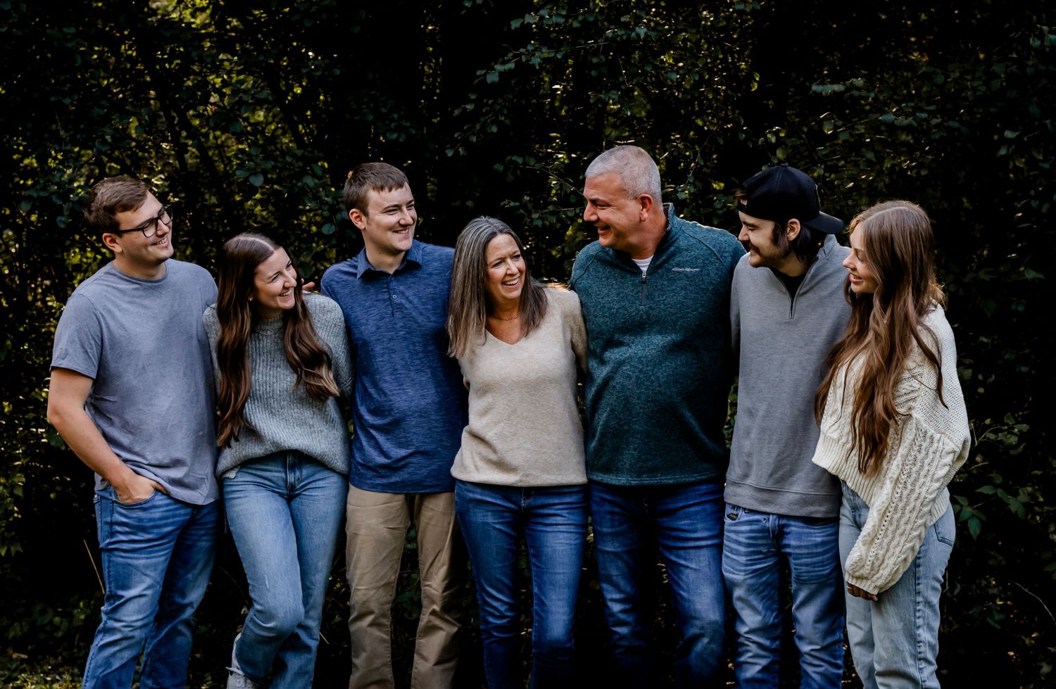 Group stands close together laughing outdoors wearing casual clothing against wooded background.