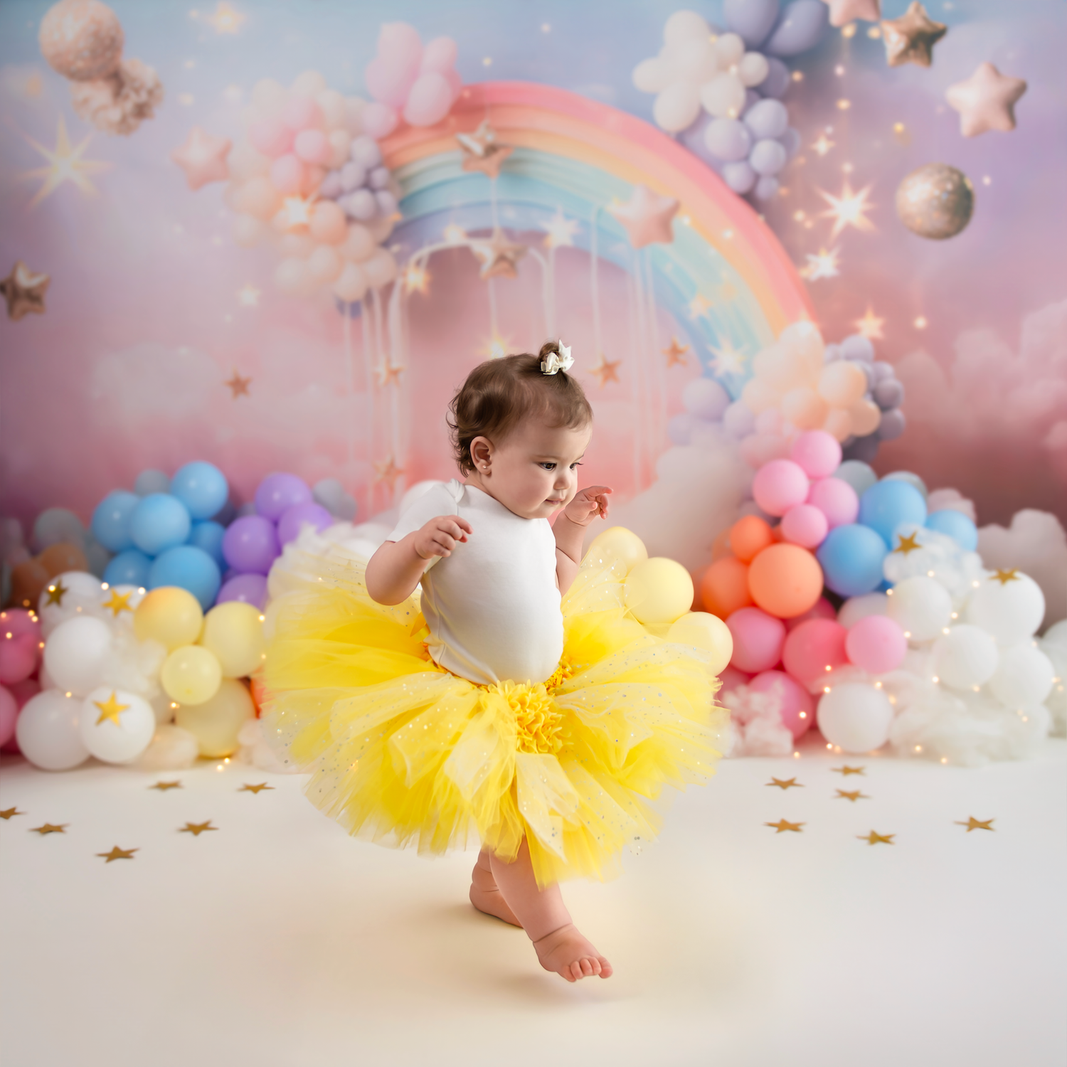 Baby in yellow tutu dress poses against dreamy backdrop with pastel balloons and twinkling star decorations.