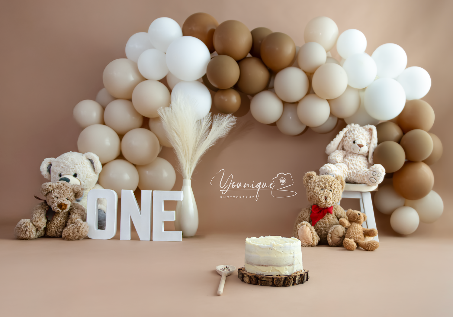 A neutral-toned cake smash setup with balloon garland, teddy bears, and 'ONE' letters for first birthday photography.