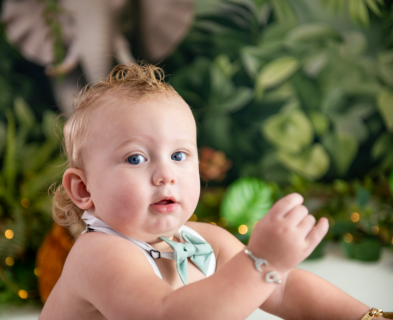 Sweet baby in a bow tie sits against a green leafy backdrop with twinkling lights.
