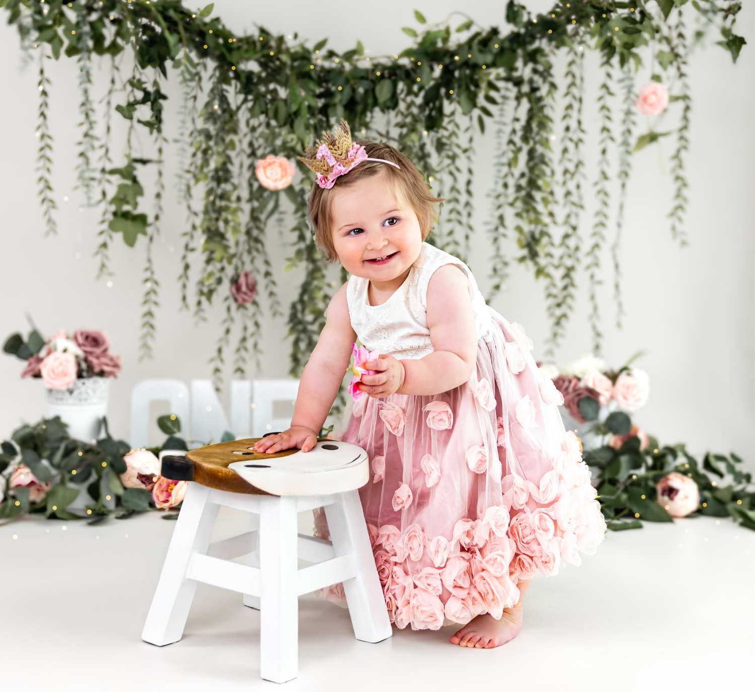 A small child in a pink ruffled dress poses near a white stool against a floral backdrop with hanging greenery.