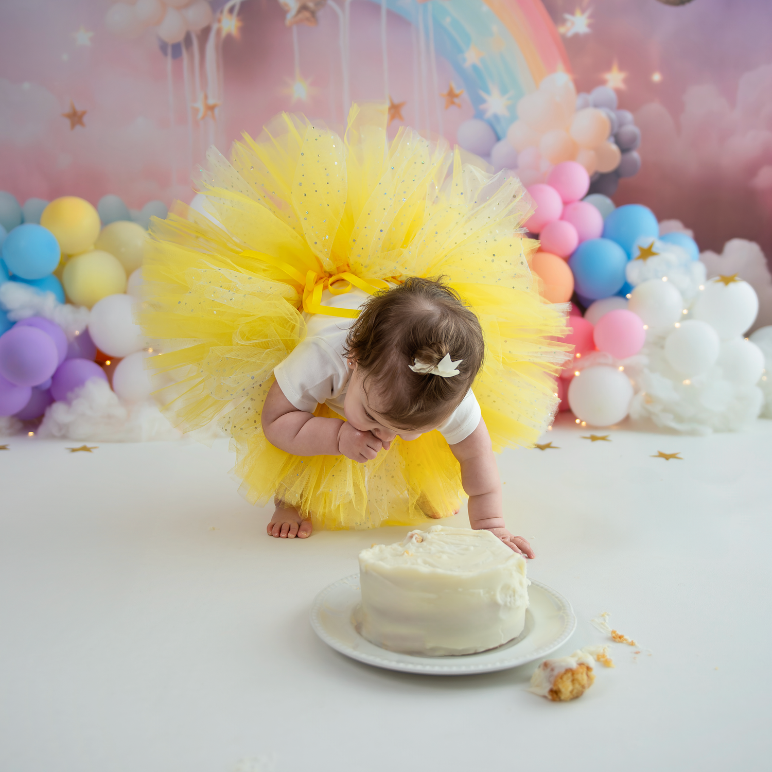 Cake smash photo session setup with yellow tulle tutu, pastel balloons, and white cake against pink backdrop.