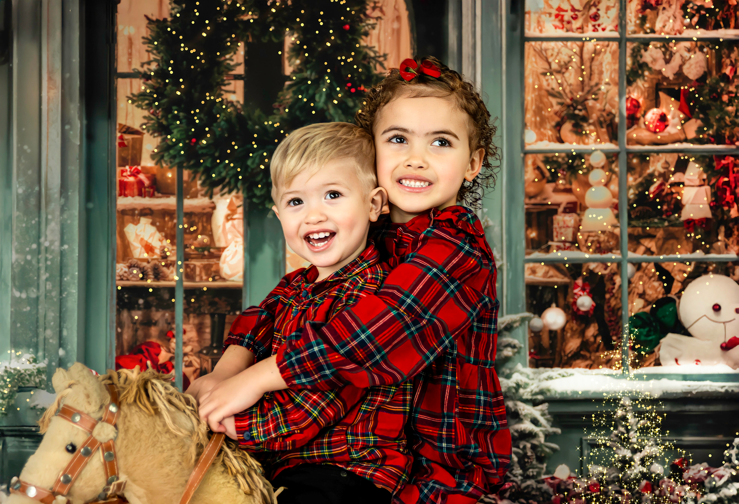 Two children wearing red plaid shirts sit by a holiday storefront display with a wreath and decorations.