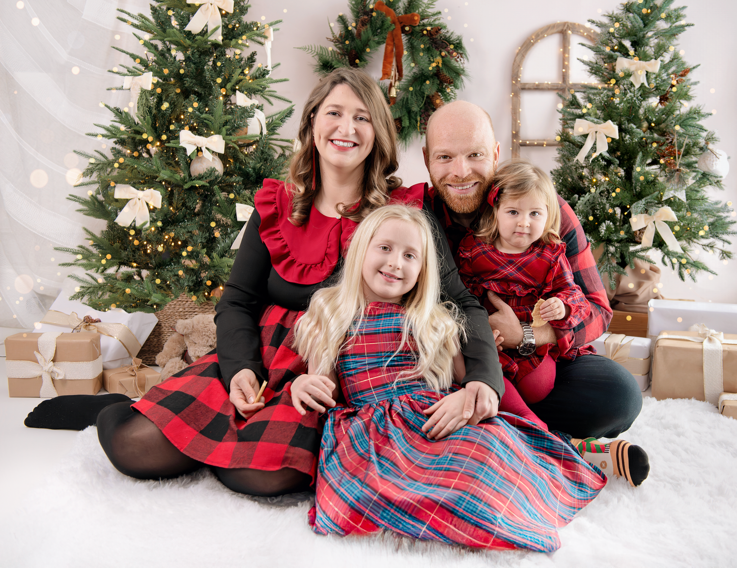 A group sits together in coordinating red and plaid holiday outfits by a Christmas tree in a bright white studio setting.