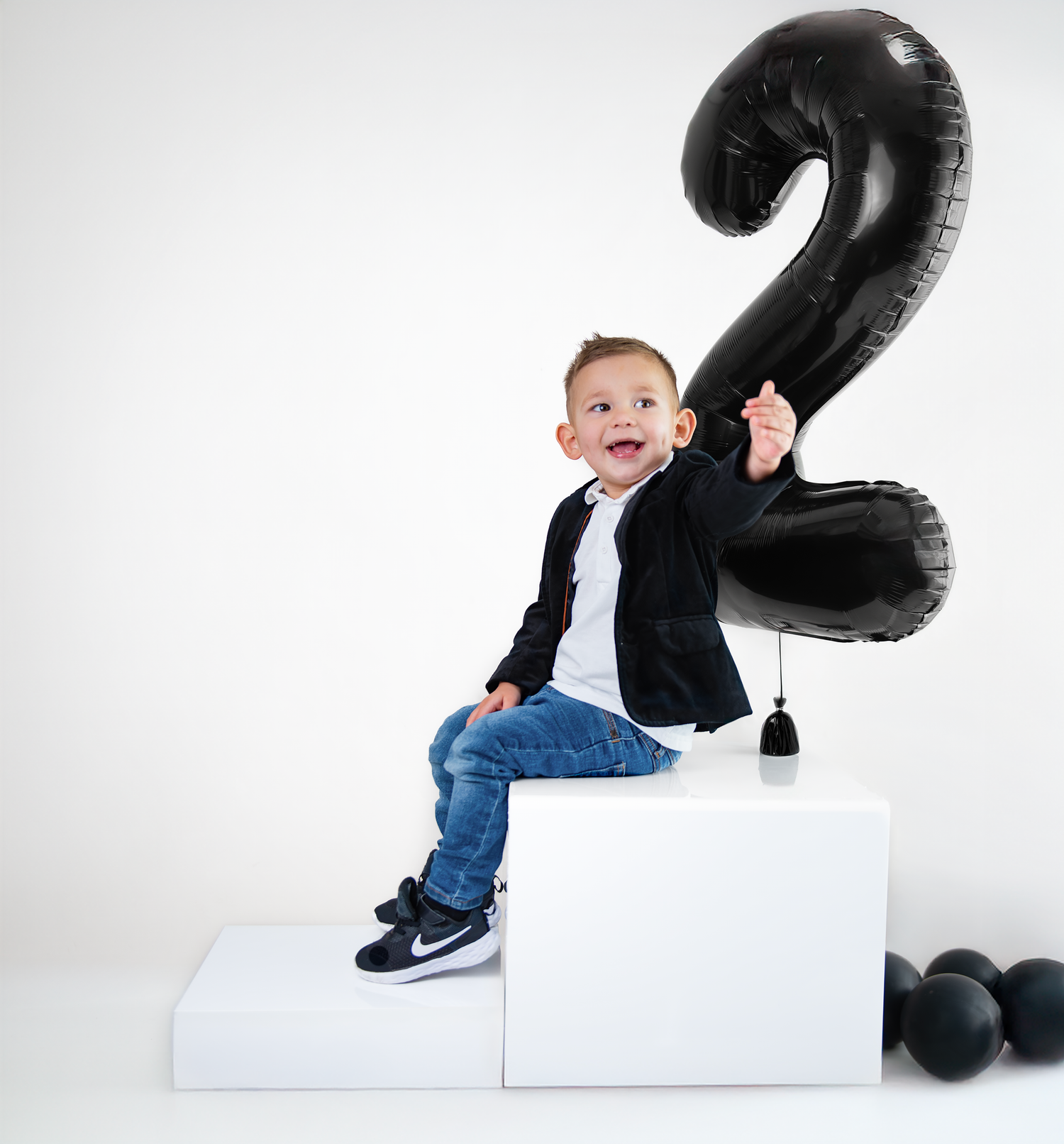 A toddler in jeans sits on white blocks next to a large black number 2 balloon for birthday photos.