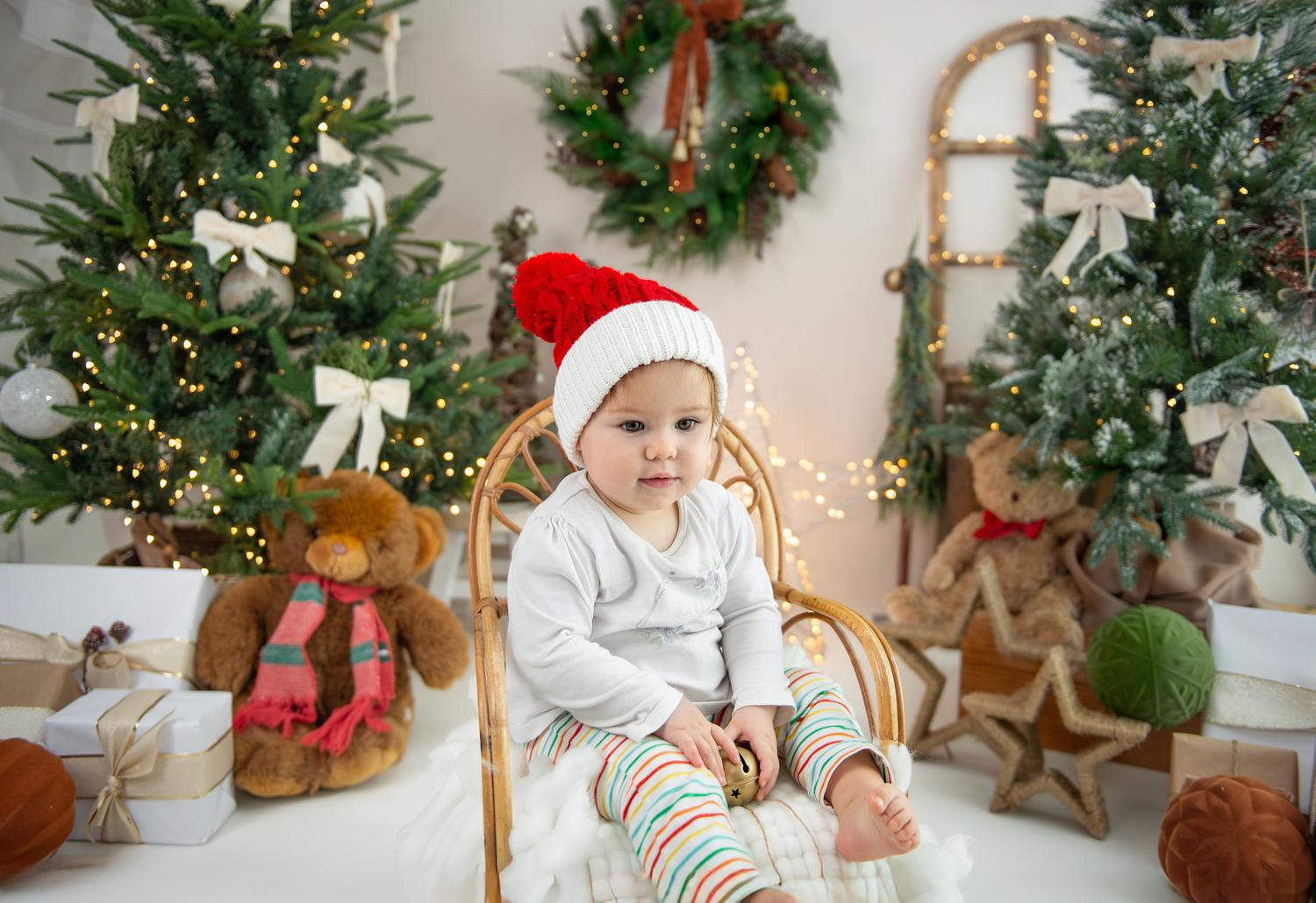 A baby in holiday attire sits on a white floor surrounded by Christmas decorations and festive greenery.