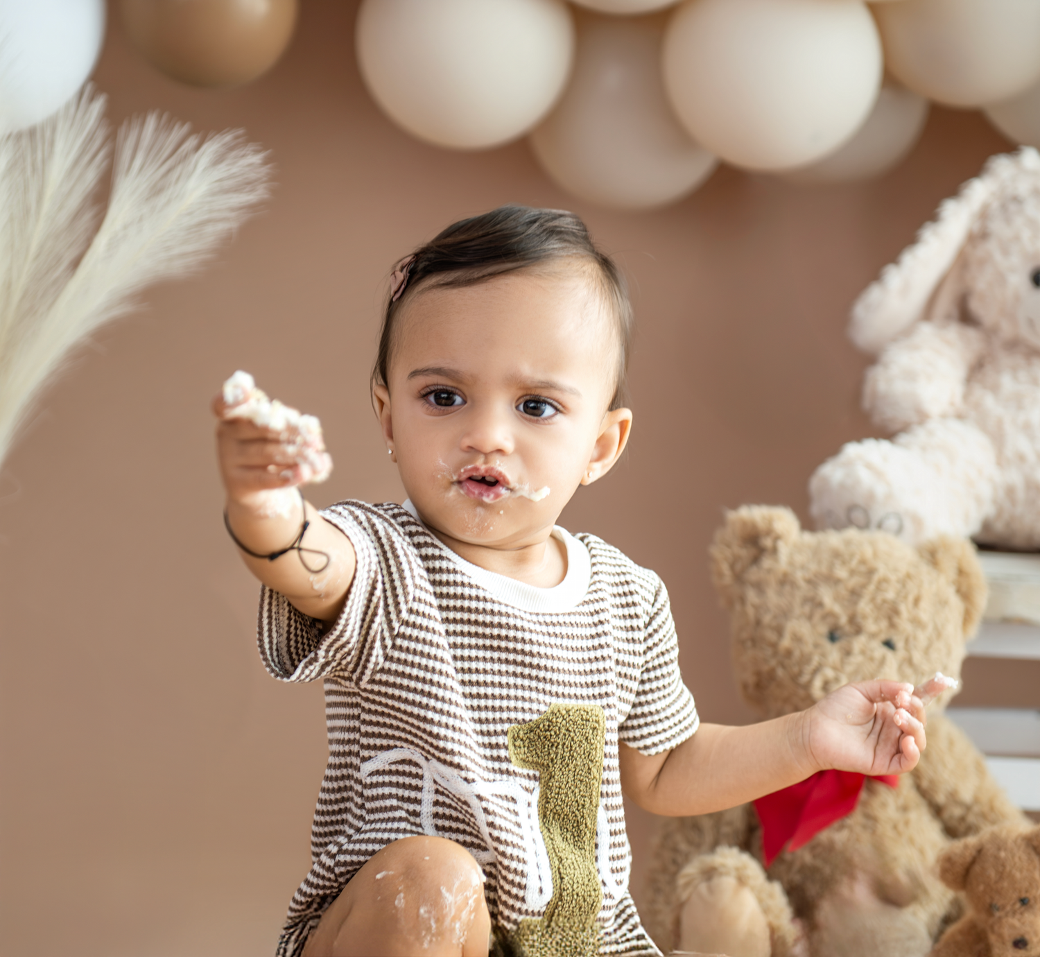Baby in brown and white striped onesie plays with white feathers and teddy bears in soft studio lighting.