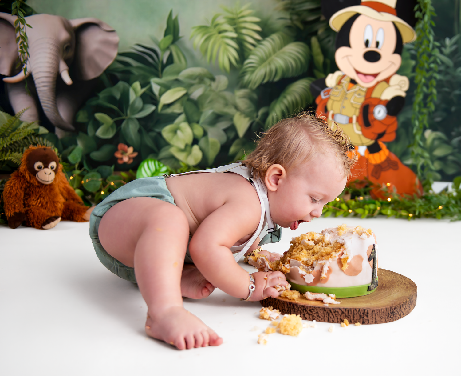 A baby crawls near a Mickey Mouse plush toy and woodland-themed decorations in a studio photo session.