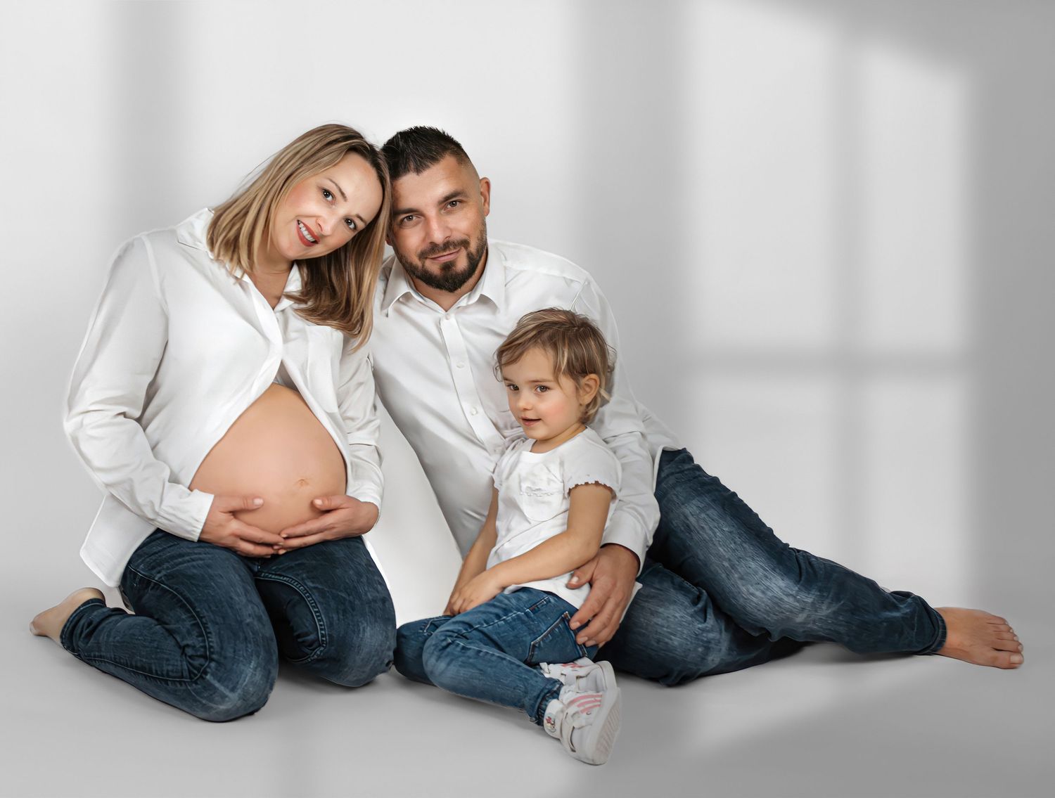 Family wearing white tops and jeans poses casually together on white backdrop in studio.
