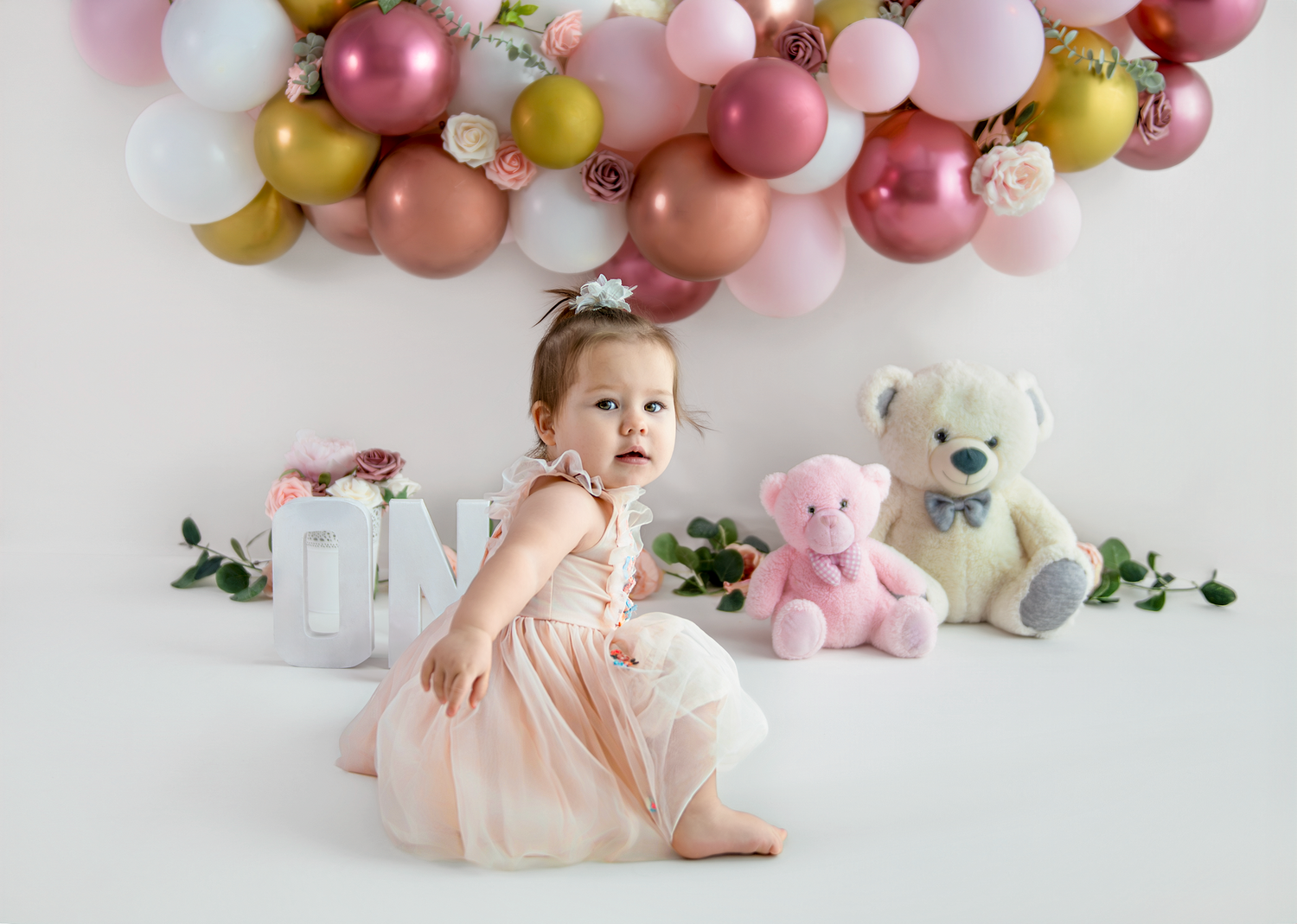 A festive first birthday setup with pink and gold balloons, stuffed animals and gifts against a white backdrop.