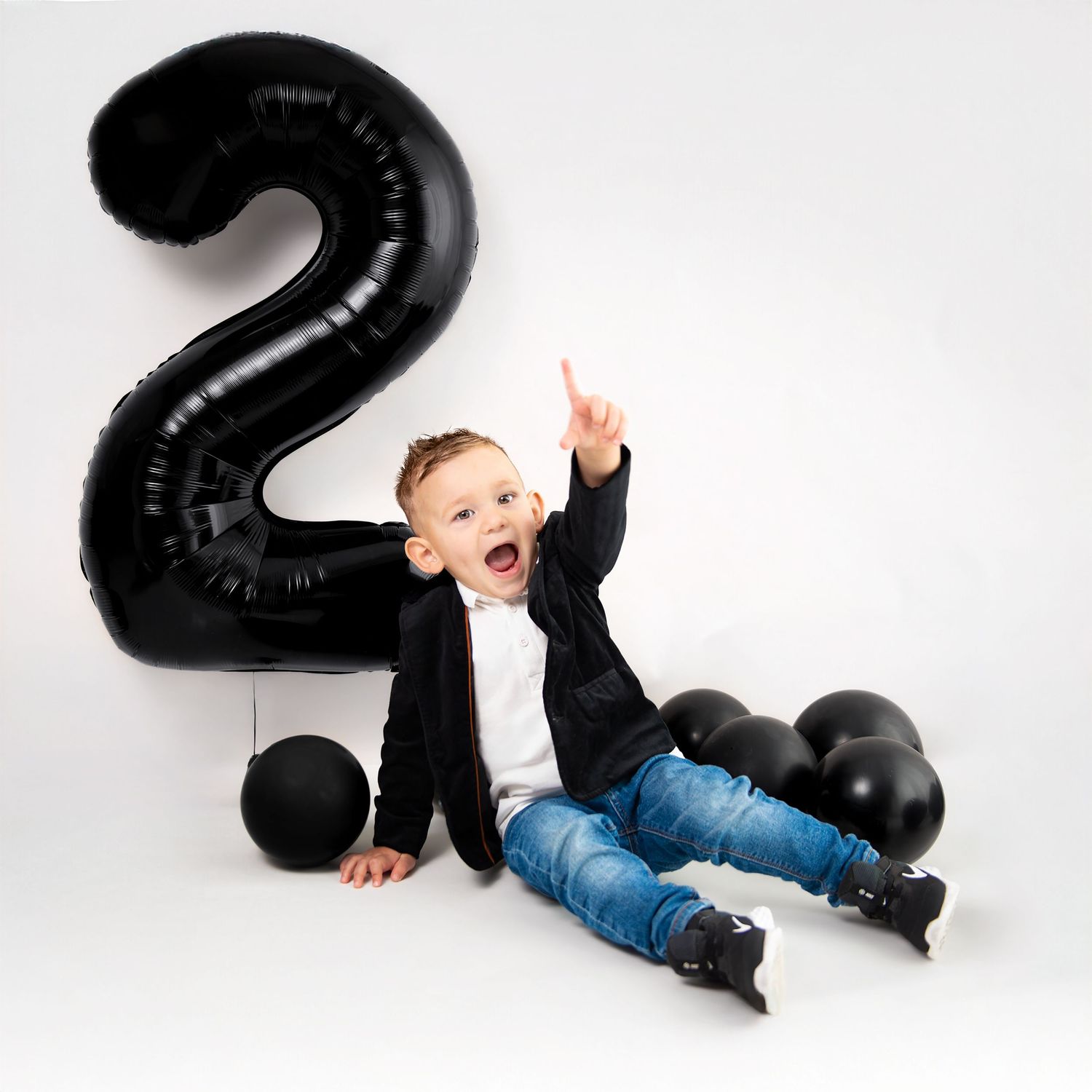 Toddler in denim and black shirt playfully interacting with large black number balloons on white background.