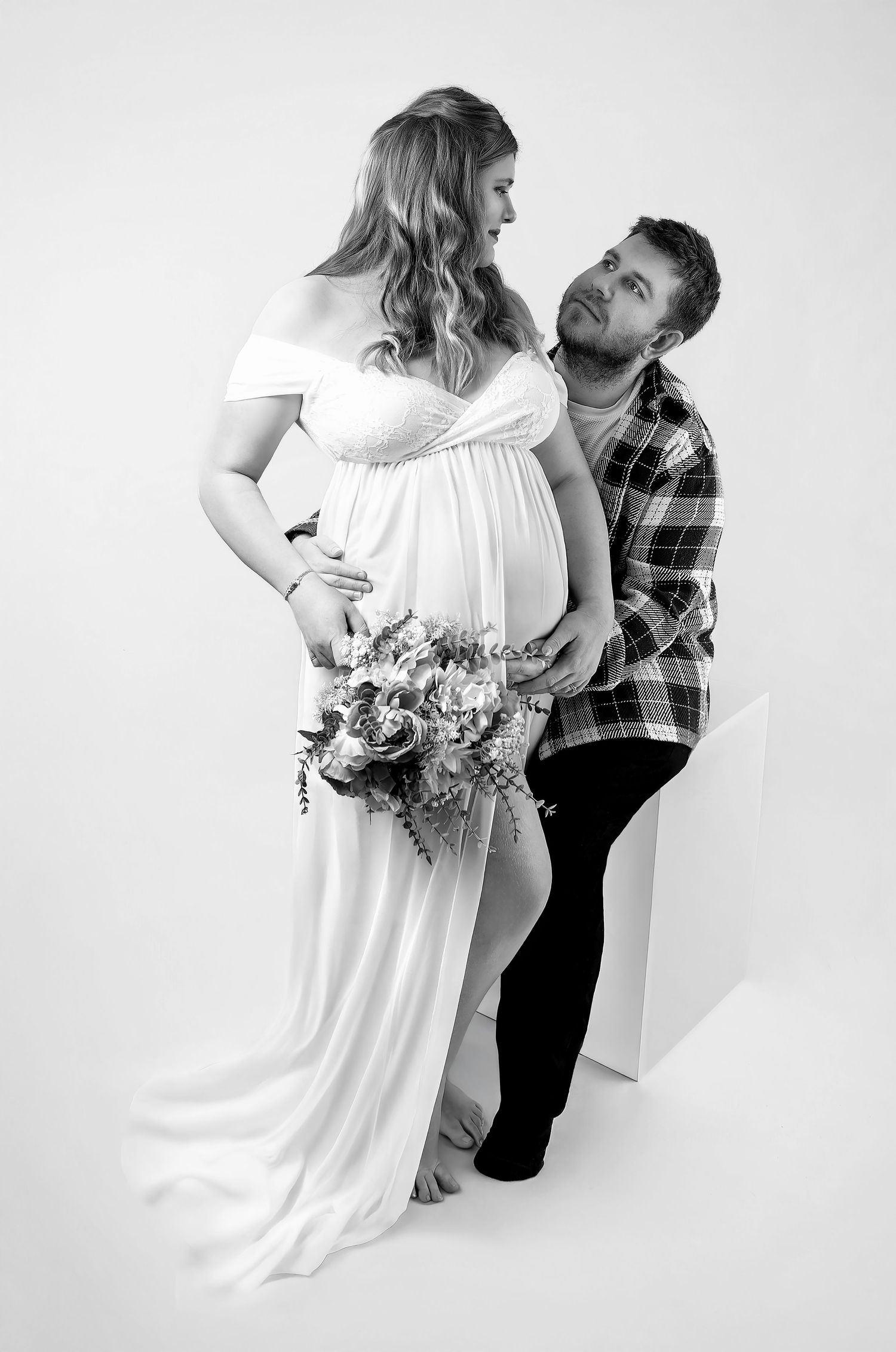 Black and white maternity photo of pregnant woman in white gown holding flowers with partner gazing at her bump.