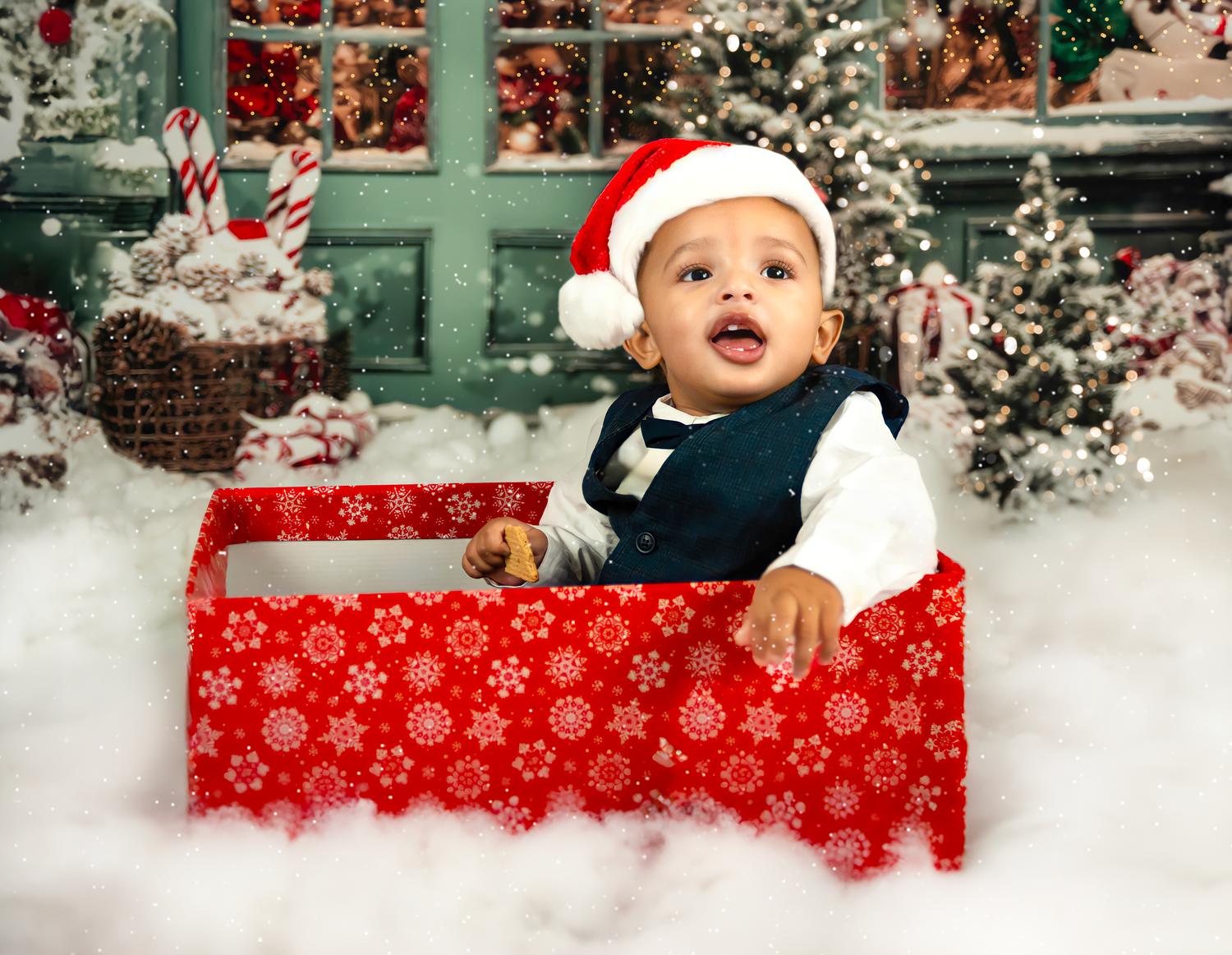 Small child in Santa hat sits in red snowflake-patterned gift box surrounded by festive Christmas decorations and snow.