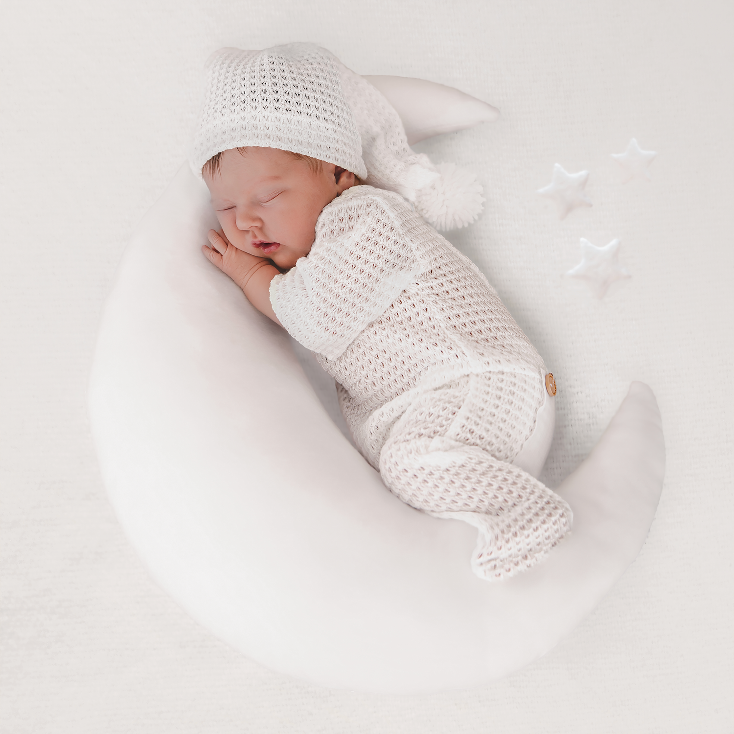 Newborn baby sleeping peacefully on white crescent moon prop, wearing white knit outfit and hat with stars.