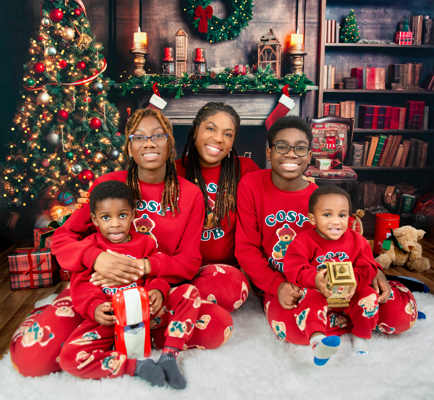 A family in matching red holiday pajamas poses together in front of a decorated Christmas tree and festive backdrop.
