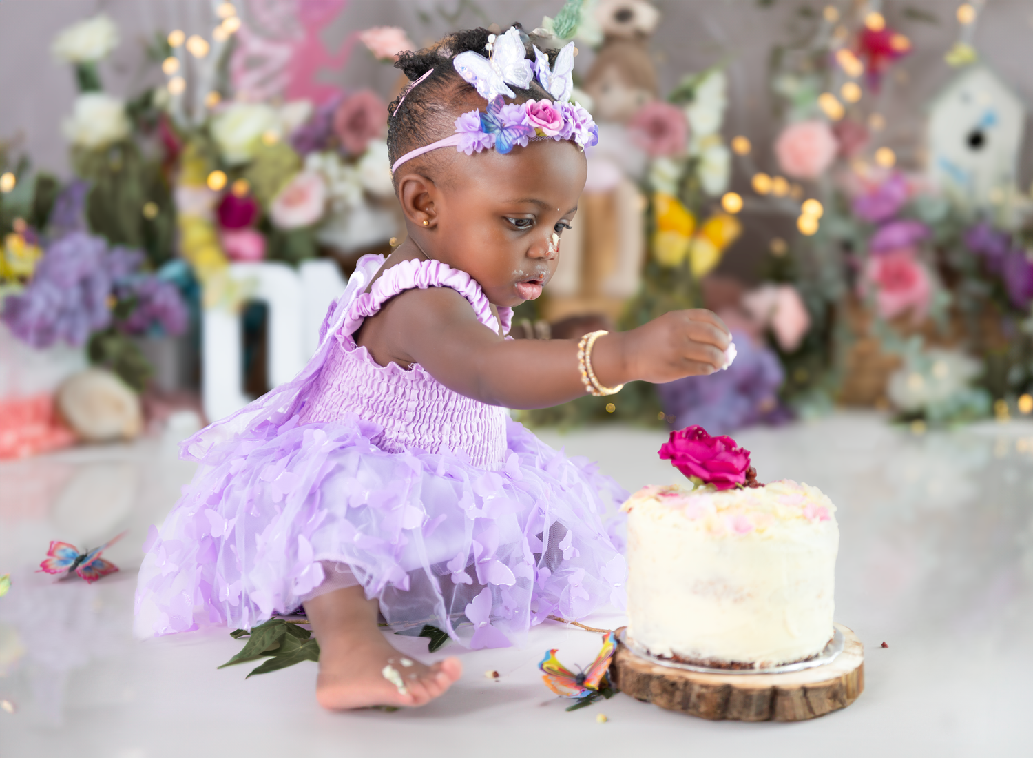 A child in a lavender tutu dress sits at a decorated table with a cake and floral decorations for a birthday celebration.
